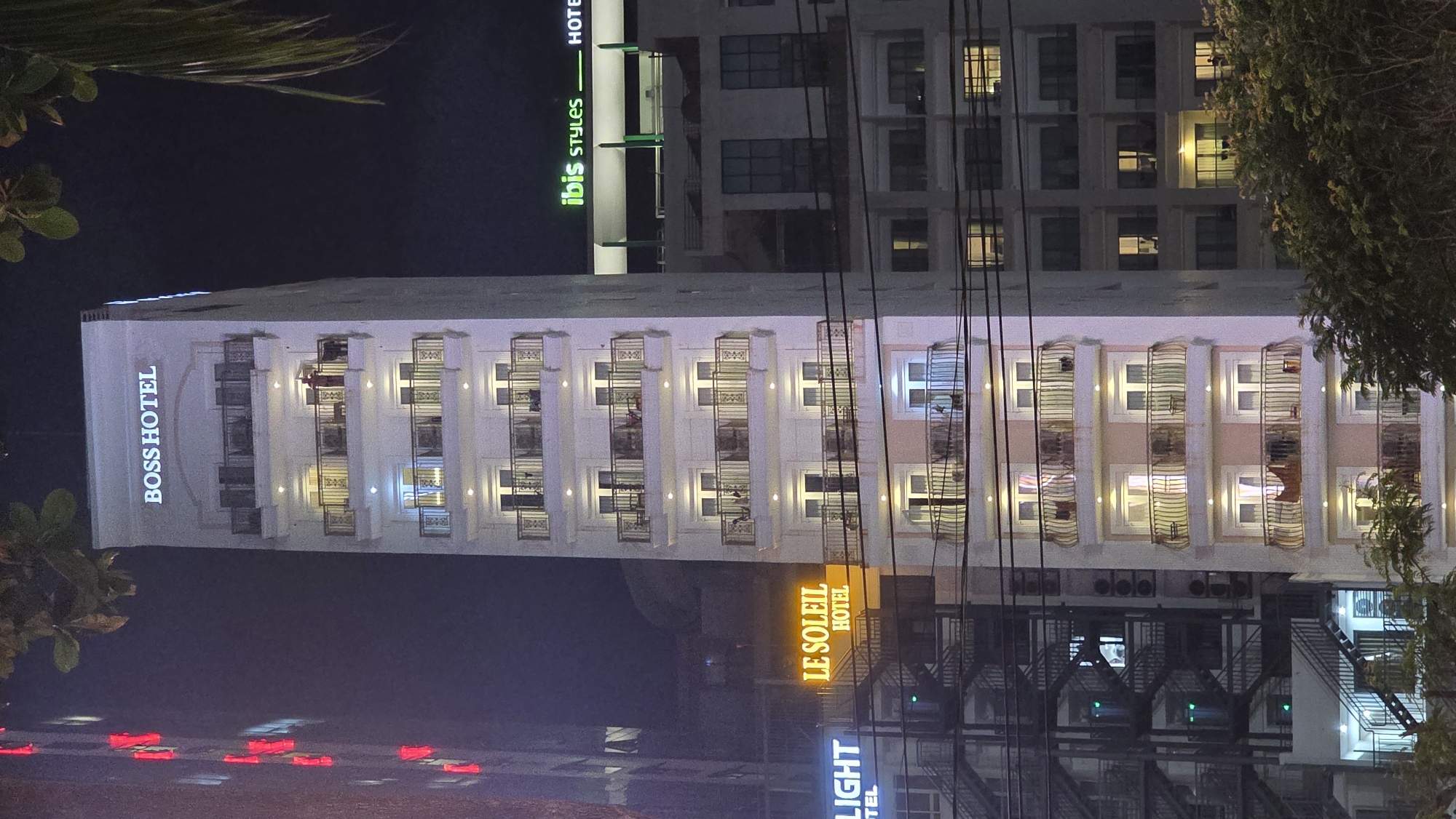 The image displays an urban nighttime scene in Nha Trang, Vietnam, dominated by several illuminated hotel buildings. The photograph is oriented vertically, showing the buildings stretching upwards.
The most prominent structure is a tall, light-colored hotel in the center, with its name "BOSS HOTEL" written vertically in large, glowing letters on its left facade. This building features multiple floors with rows of windows, many of which are lit, revealing interior activity. Architectural details like vertical columns or pilasters divide the window sections.
To the right and slightly behind the "BOSS HOTEL," another hotel with a distinctive green and white sign is visible, reading "ibis STYLES HOTEL." This building appears taller and has a grid-like pattern of windows, some also illuminated. Further to the right, tangled power lines or utility cables cross the frame, partially obscuring the view of the buildings.
Towards the lower right, an illuminated sign prominently displays "LE SOLEIL HOTEL" in bright yellow and orange lettering. Below this, a partial sign for "LIGHT HOTEL" can be seen. This area also shows parts of building facades and lit windows.
The time of day is clearly night, indicated by the dark sky and the reliance on artificial lighting from the hotels and their signs. Dark foliage from trees is visible on the far left and right edges of the frame, contrasting with the man-made structures. The bottom left corner shows blurred red lights reflecting, possibly from street-level signs or vehicles. The overall scene suggests a busy, well-lit hotel district.