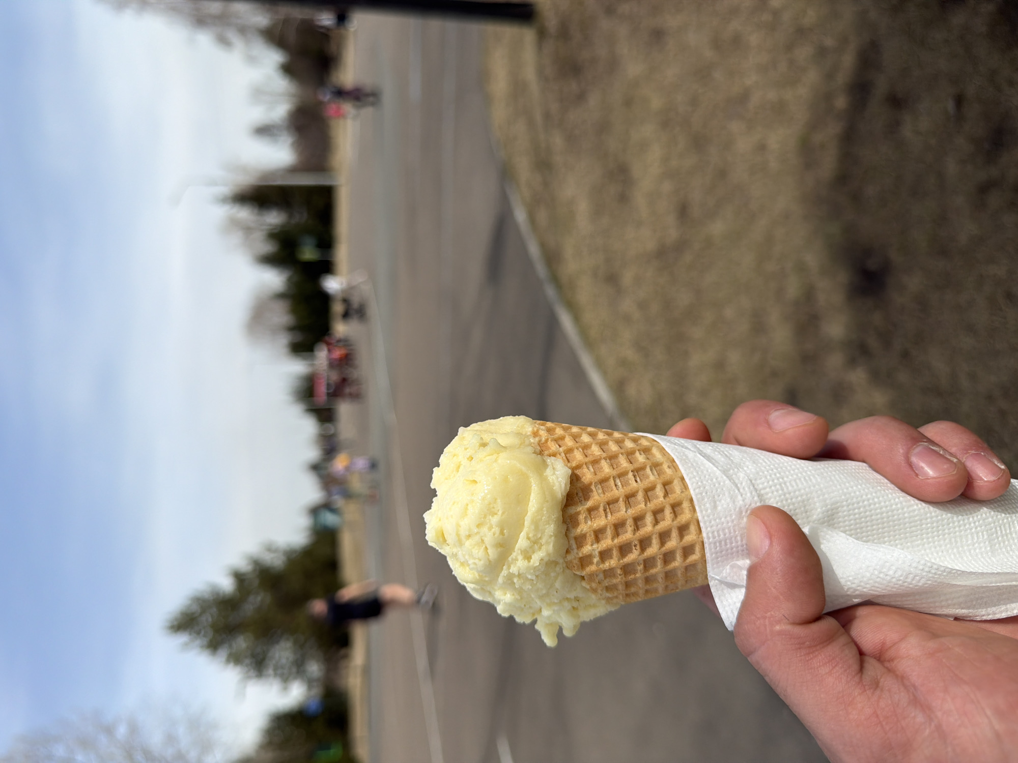 A person is holding an ice cream cone with yellow ice cream in their right hand, wrapped in a white napkin. The background shows a paved area with blurred figures of people and trees. The setting could be a park or recreational area. Based on the landscape, the image is possibly taken in Krasnoyarsk, Russia.