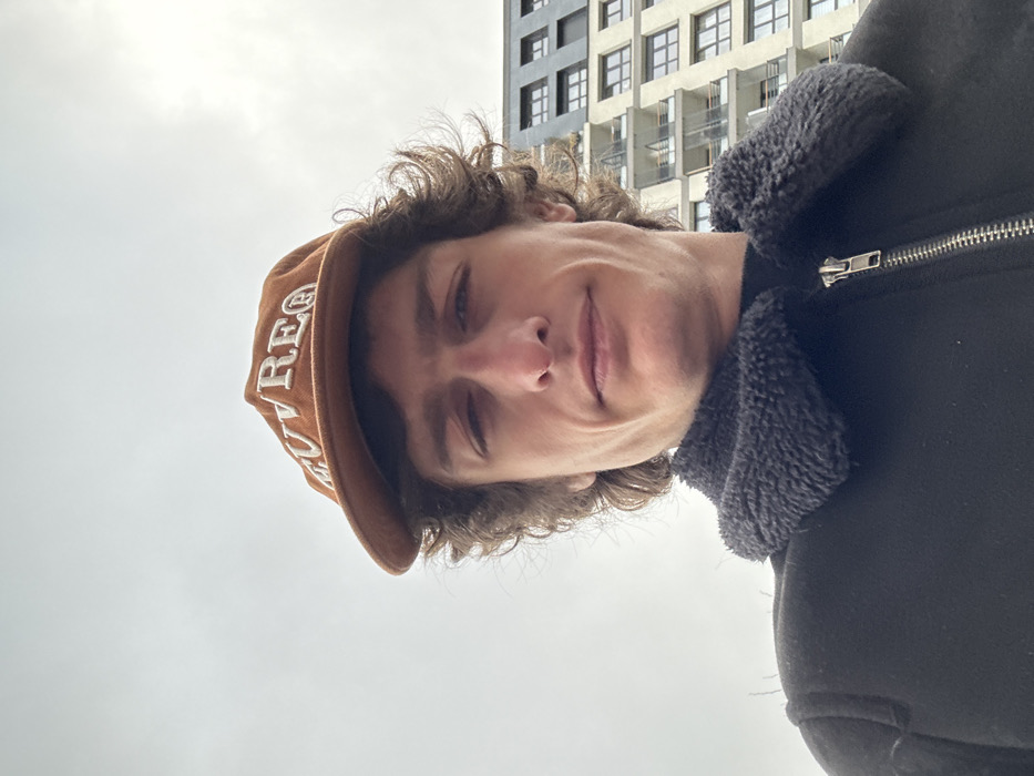 A male individual is depicted in a close-up, upward-angled selfie shot, positioned outdoors in Joinville-le-Pont, France. He appears to be in his late 20s or early 30s, with a slender face, prominent jawline, and curly brown hair visible beneath his headwear. His expression is neutral, with a slight upturn at the corners of his mouth, and he is looking directly at the camera.
He is wearing a brown baseball cap with the word "MUVREG" (or similar lettering) embroidered in cream-colored capital letters across the front. His attire includes a black jacket featuring a dark grey or black fleece/shearling collar and a visible silver-toned zipper pull on the front. A dark-colored garment, possibly a turtleneck, is visible beneath the jacket.
The background on the left consists of a uniformly overcast, light grey sky. To the right, a section of a multi-story modern building is visible, characterized by light-colored exterior panels, numerous dark-framed windows, and several balconies with transparent or semi-transparent railings. The architecture suggests a contemporary residential or office structure. No other specific ground-level features or landmarks of Joinville-le-Pont are discernible.