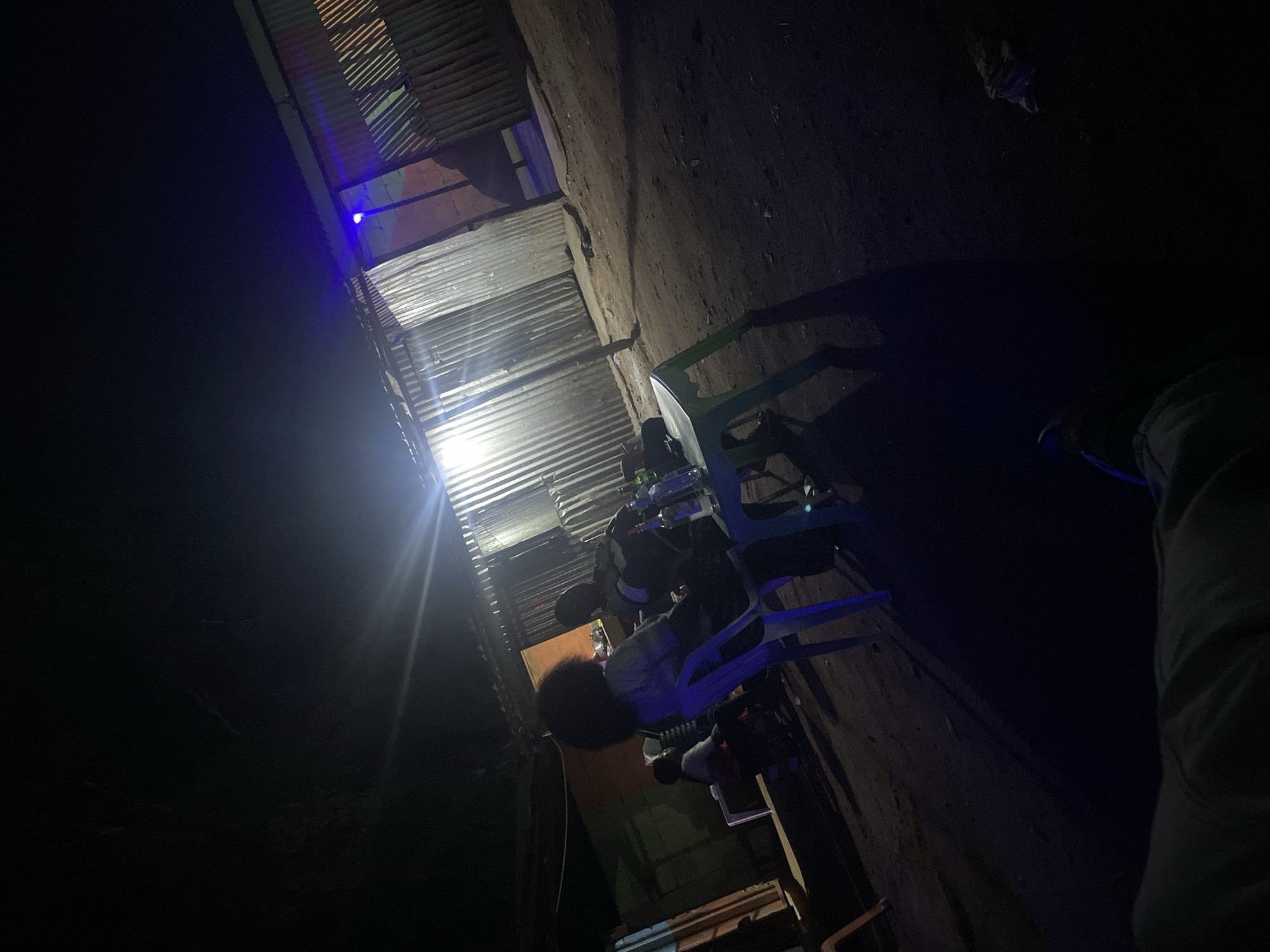 A low-angle, poorly lit photograph shows a person seated on a blue plastic chair under a corrugated metal roof in Jos, Nigeria. The person has dark hair and is wearing a light-colored top and dark pants. They appear to be facing away from the camera. The chair is positioned in a narrow space between the corrugated metal structure and a rough, earthen wall. The lighting is dim, with a bright light source emanating from within the structure, casting long shadows. The surrounding environment appears to be dark and possibly enclosed. The scene evokes a sense of urban poverty and informal living conditions.