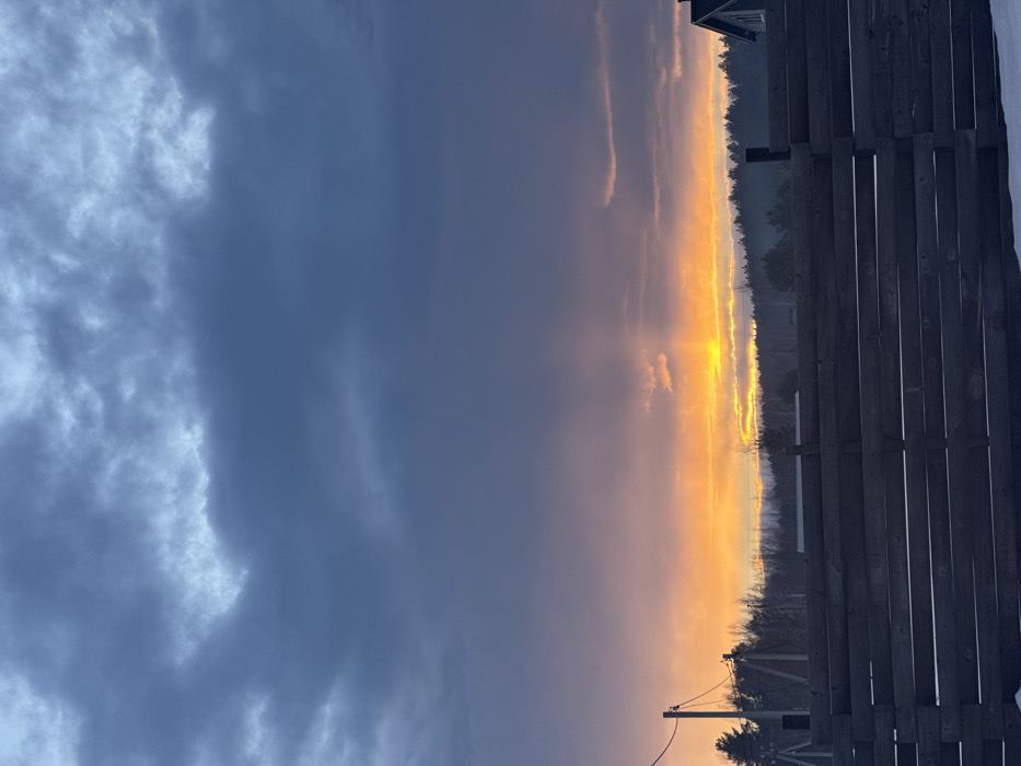 The image captures an outdoor scene in Kamskoe, Russia, during a sunset or sunrise. The sky dominates the upper two-thirds of the frame, characterized by dense, dark blue-grey clouds overhead transitioning to a bright orange and yellow glow near the horizon, from which distinct sunbeams emanate. A dark, horizontally-slatted wooden fence occupies the lower third of the image, running parallel to the viewer. Below the fence, a thin layer of white snow is visible on the ground. A utility pole with wires is positioned on the left side, extending upwards from behind the fence line. In the mid-ground, a dark, dense treeline or forest is silhouetted against the vibrant sky. On the far right, a portion of a building with a dark roof is visible above the fence. No individuals, actions, or interactions are present.