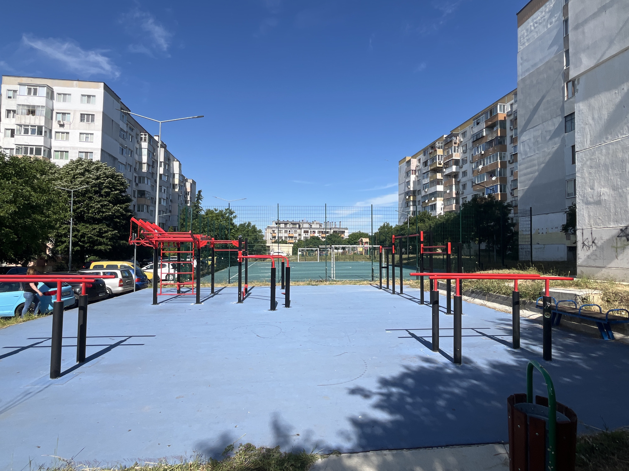 This is a photograph of an outdoor exercise park located in the city of Varna, Bulgaria. The park features a bright blue rubberized surface with various pieces of red and black exercise equipment, including parallel bars, pull-up bars, and monkey bars. In the background, there is a fenced sports field with a goal, and beyond that, several large apartment buildings characteristic of Soviet-era architecture. A few cars are parked along the street adjacent to the park. The sky is clear and blue with a few scattered clouds. The overall impression is one of a functional public space designed for recreation and fitness within an urban residential area.
