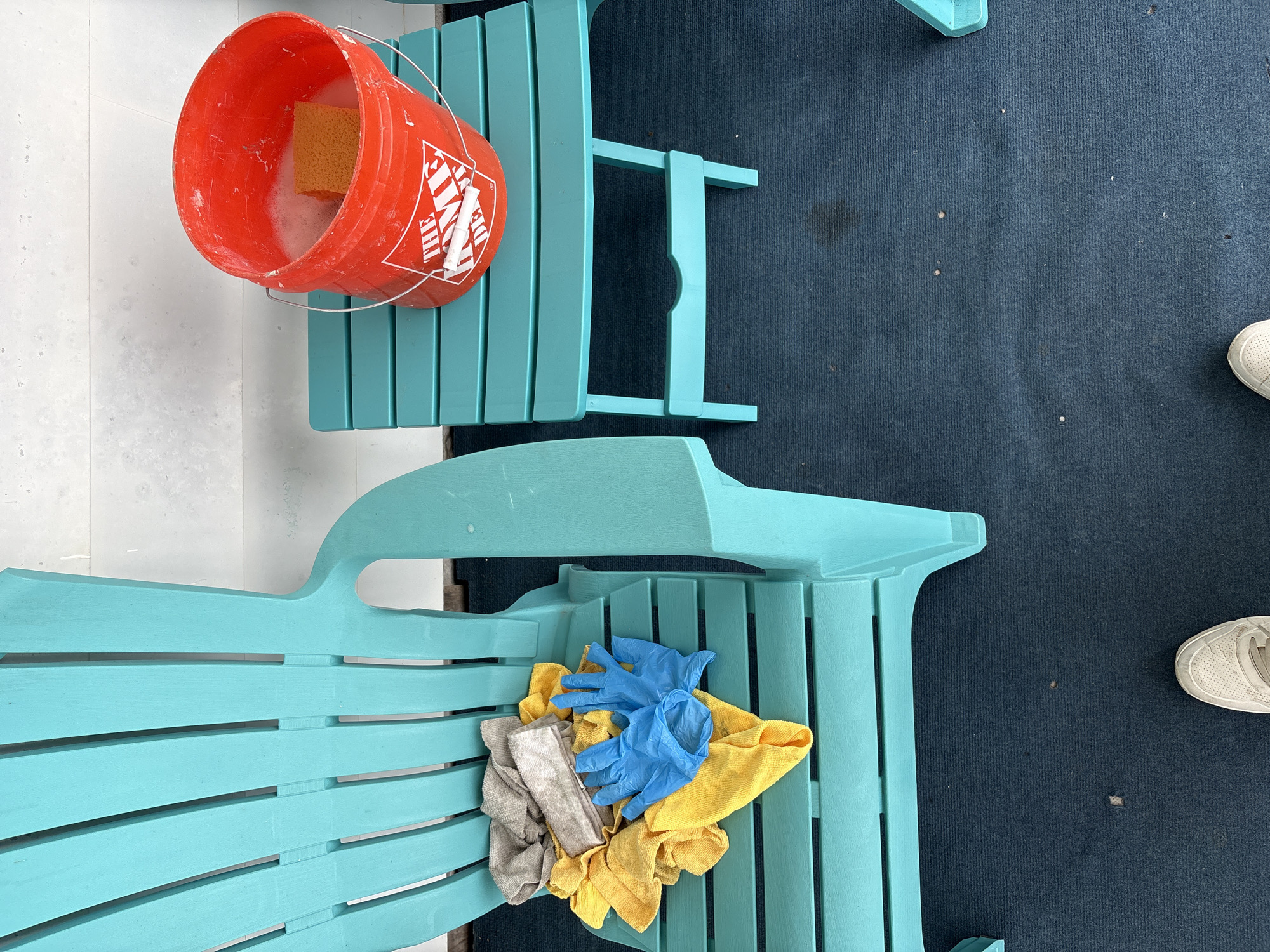 This media file captures an overhead view of an outdoor cleaning setup, likely on a patio or deck in Hampton, USA.
The main subjects are two teal-colored Adirondack chairs. The top chair has a bright orange Home Depot bucket resting on its side, filled with soapy water and a yellow sponge. The bucket prominently displays "THE HOME DEPOT" logo. The bottom chair holds a pile of cleaning supplies, including a pair of blue rubber gloves, several yellow microfiber cloths, and a gray cleaning rag.
The scene suggests an activity of cleaning the outdoor furniture. The presence of the bucket with soapy water, sponge, gloves, and cloths all indicate that the chairs are either being cleaned or have just been cleaned.
Notable details include the ground surfaces: a light-colored, possibly white, tiled or paved area on the left, and a dark blue outdoor carpet or mat covering the right side. A person's foot, wearing a white sneaker, is partially visible on the carpet in the bottom right corner of the frame. The lighting appears bright and even, characteristic of daytime.