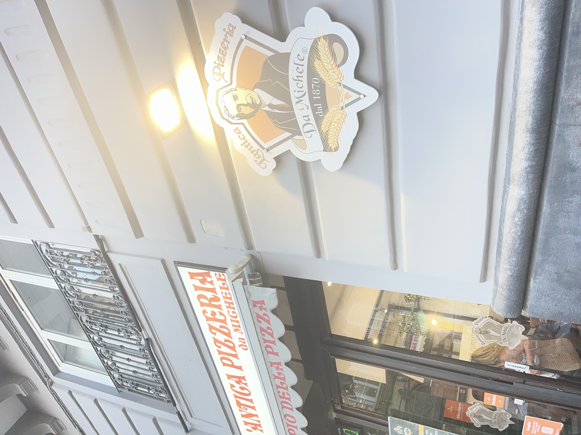 The image shows the facade of an "Antica Pizzeria da Michele" in Naples, Italy. A prominent orange sign with white lettering reads "ANTICA PIZZERIA da MICHELE" above a red scalloped awning that says "L'IMPERIO DELLA PIZZA" in white lettering. To the right, a round emblem-like sign features a portrait of a man in a suit and tie, with the text "l'Antica Pizzeria Da Michele dal 1870" surrounding him. Wheat stalks are depicted below the portrait. The building has a grey, horizontally striped facade. A balcony with decorative metal railings is visible on the upper floor. The entrance area is partially visible through the glass windows, showing people and interior details. The scene is brightly lit by sunlight and an overhead light fixture.