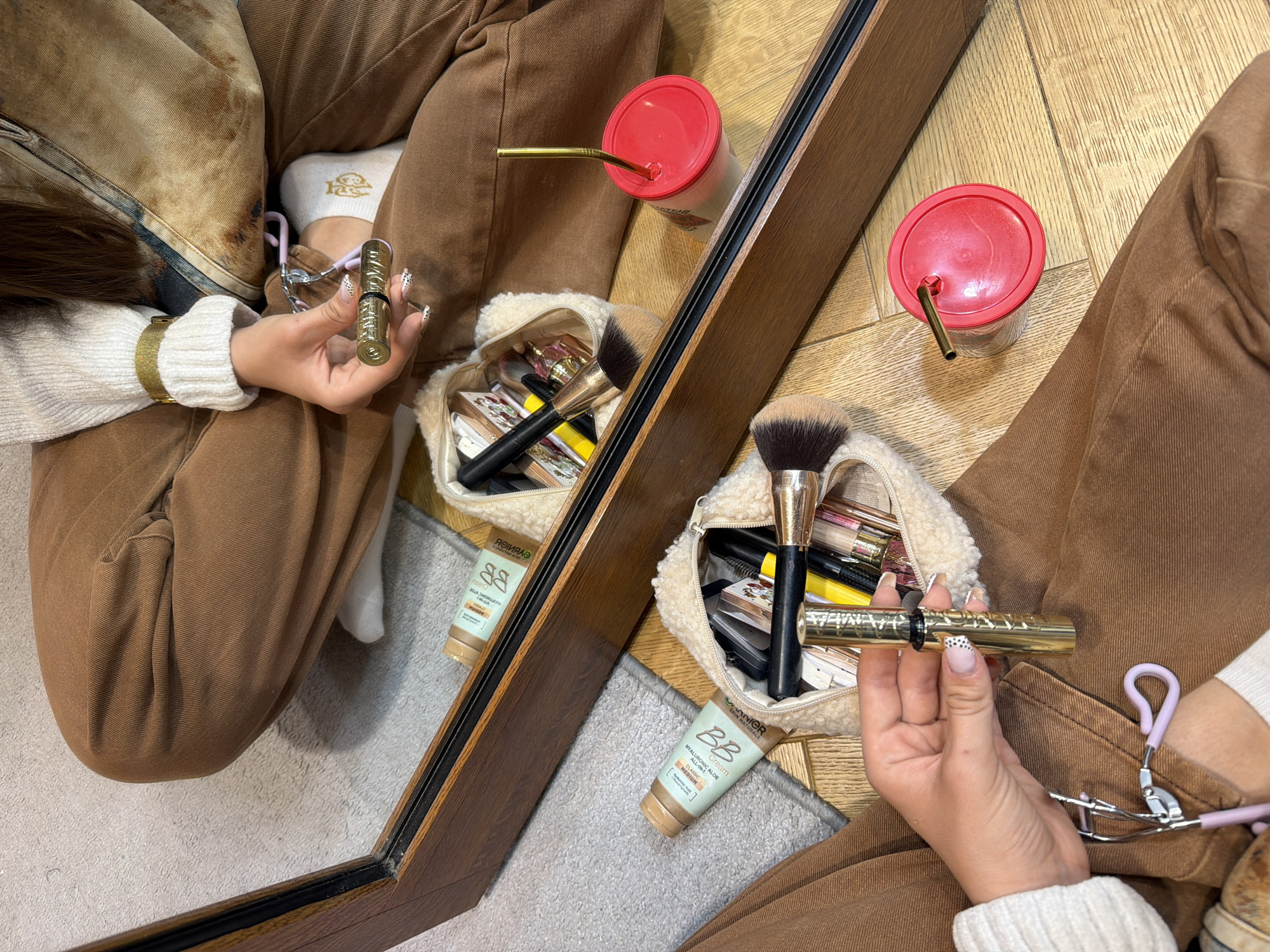 The image captures a person, likely female, sitting on the floor in an indoor setting in Buhovo, Bulgaria, reflected in a large mirror with a dark wooden frame. The person is wearing brown corduroy-style pants, a white ribbed sweater, and a white sock adorned with a small cartoon character. Their hands are visible; one holds a gold-colored mascara tube, and a lavender eyelash curler rests near their other hand/leg. Their nails feature a white and black spotted design.
Beside the person is a fluffy beige makeup bag overflowing with various cosmetic items, including several makeup brushes and eyeshadow palettes. A tube of "GARNIER SKIN NATURALS BB HYALURONIC ALOE ALL-IN-1 CLASSIC Cream MEDIUM" is prominently displayed on the floor next to the mirror. A red plastic cup with a gold reusable straw is also visible, resting on a light wooden floor section, part of which is reflected in the mirror. A light grey rug or carpet covers another part of the floor. The scene depicts a moment of personal grooming or makeup organization.