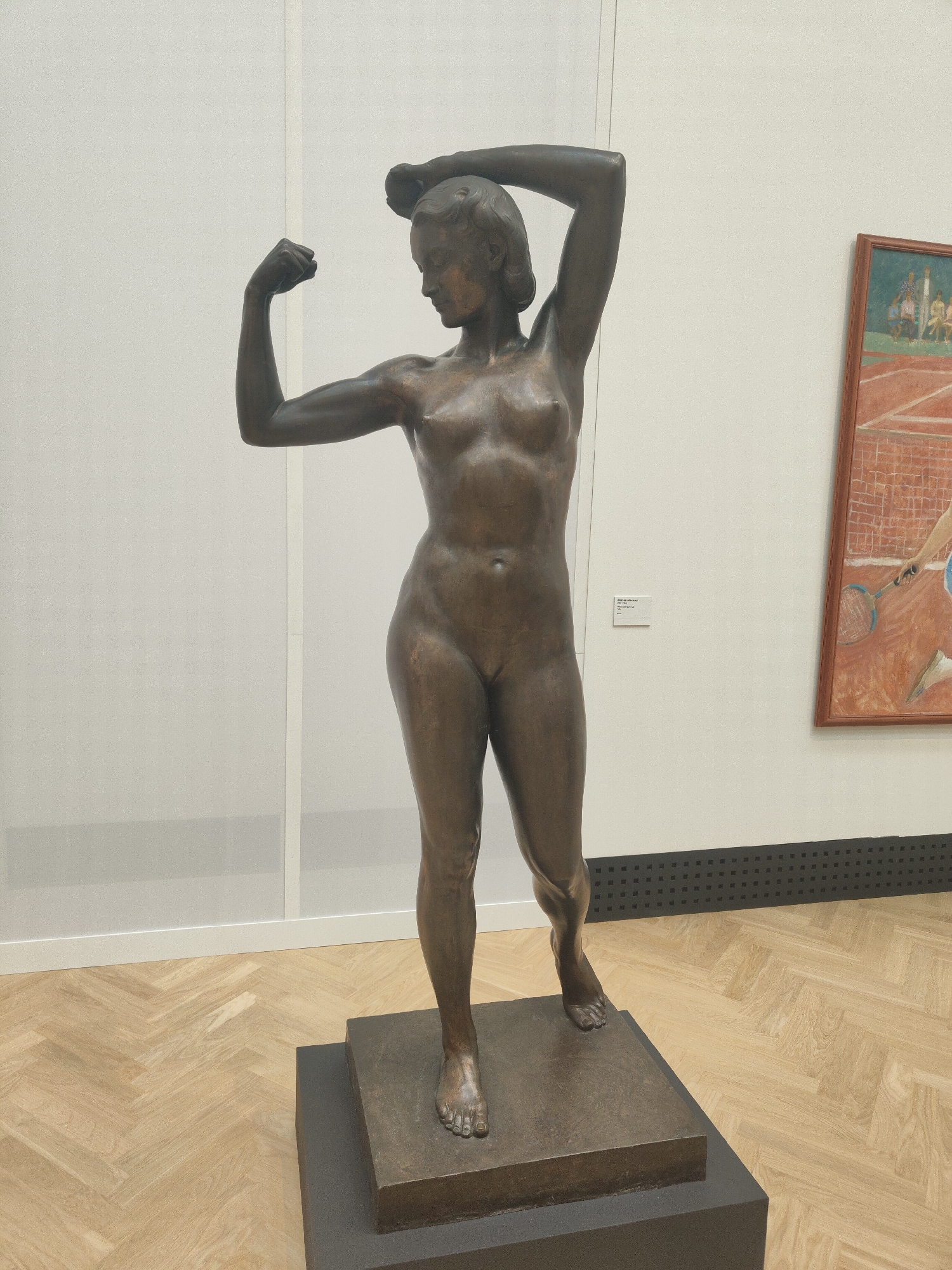 The image shows a bronze sculpture of a nude female figure on a tiered base in what appears to be a museum or gallery setting. The woman is depicted with one arm raised and bent at the elbow, fist clenched, while the other arm is raised and her hand rests on her head. The sculpture has a dark bronze patina. In the background, there is a white wall and a framed painting depicting a tennis scene. The floor is made of light-colored parquet. A small sign or label is affixed to the wall next to the sculpture.