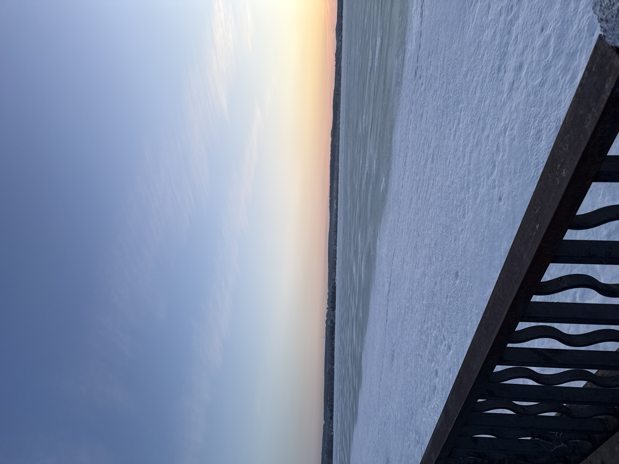 The image depicts a winter scene, possibly a frozen lake or river, viewed from a high vantage point, likely a bridge or embankment. A wrought iron railing is visible in the foreground, suggesting the location is a public walkway. The sky transitions from blue to a soft orange-yellow hue, indicating either sunrise or sunset. The surface of the frozen expanse is covered in snow or ice. Given the prompt context, it is presumed this image was taken in Votkinsk, Russia, possibly along the shoreline of the Votkinsk Reservoir or another body of water within the city. No people are visible. The scene is static and peaceful.