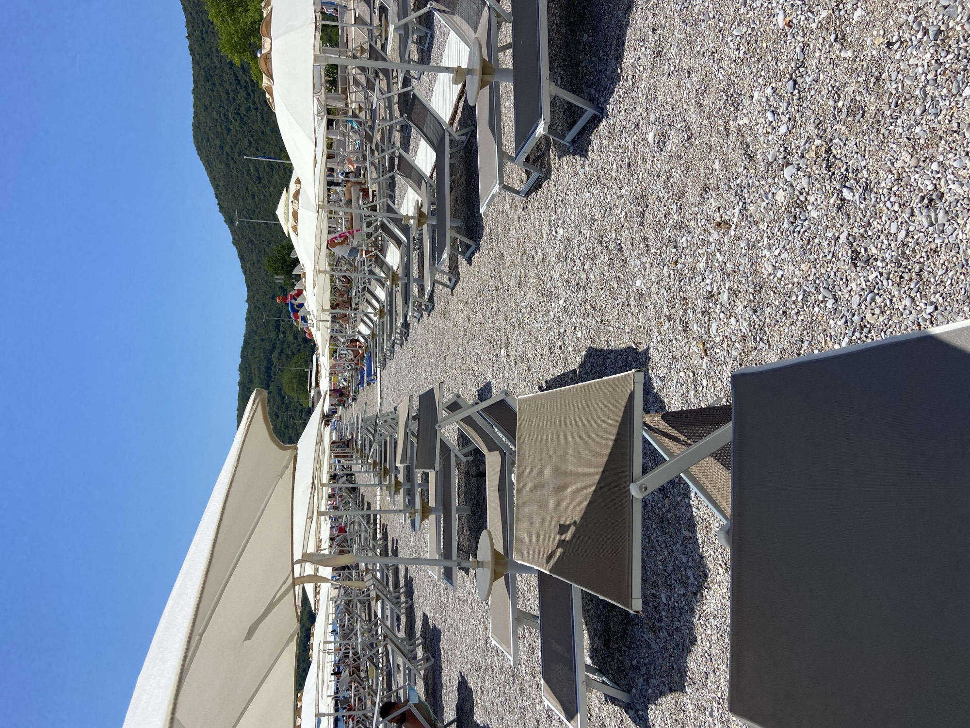 The image depicts a sun-drenched beach scene in Prijevor, Montenegro. Rows of empty lounge chairs and umbrellas stretch along a pebble-strewn shore, suggesting a popular tourist destination. The sun is high in the sky, casting sharp shadows and indicating it is likely midday. The water is a vibrant blue, hinting at a clear and pleasant day. In the background, a lush, green mountain slopes down towards the sea, adding a scenic backdrop to the resort-like setting. While most chairs are unoccupied, a few people can be seen scattered among them, relaxing or enjoying the beach. One person is dressed in what appears to be a Spider-Man costume, adding a playful element to the scene. The overall atmosphere is one of leisure and relaxation, typical of a beach holiday. There is no visible text in the image.