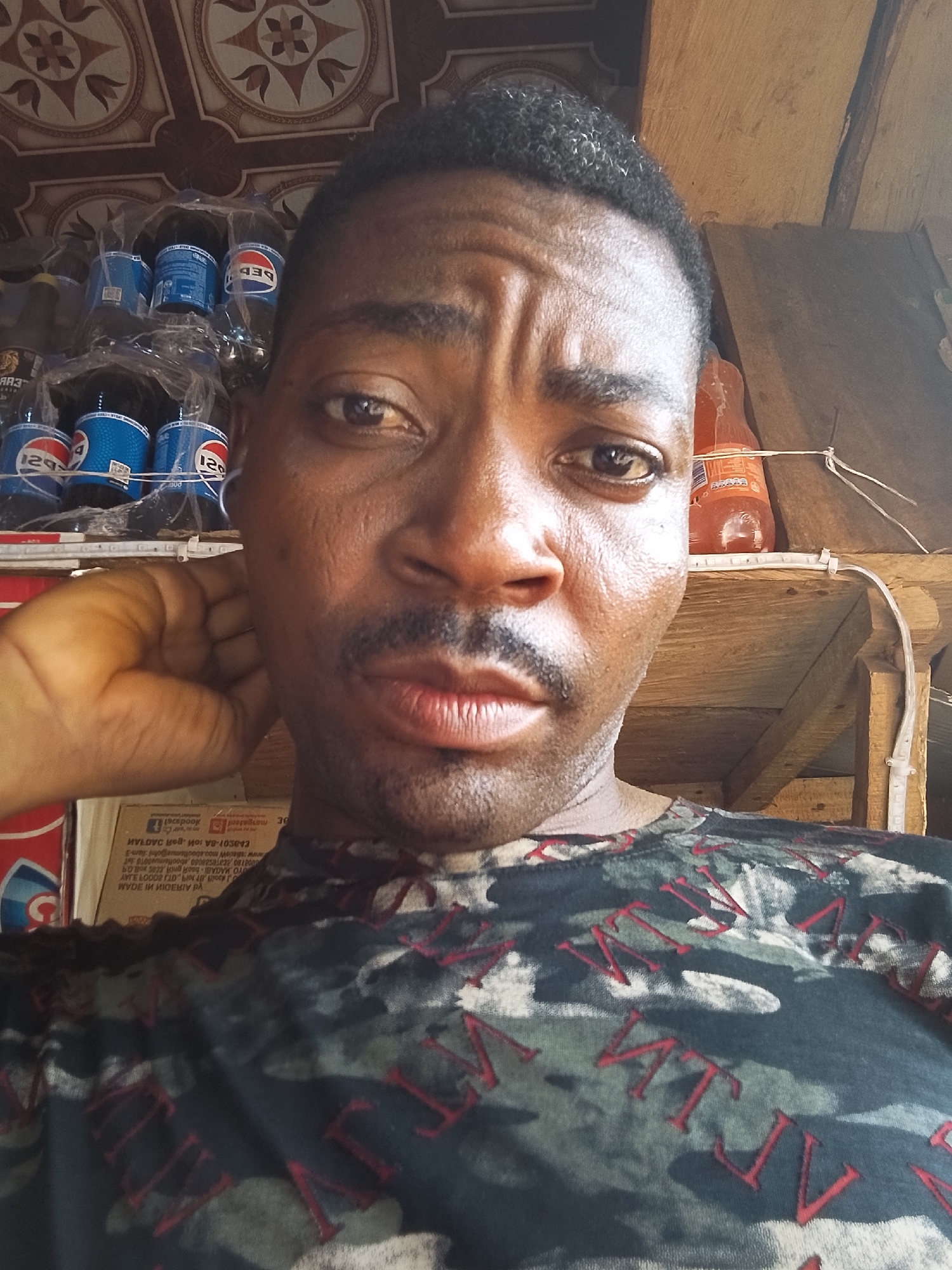 A close-up shot depicts a dark-skinned man with a mustache, wearing a camouflage-patterned shirt, likely in a small shop in Ejule, Nigeria. Behind him are shelves stocked with drinks, including Pepsi. The setting appears to be a local business. Part of a sign is visible with some legible writing, with a URL that contains "elladowmoo.aboollsmus". He is looking at the camera with a slight frown and his left hand is up to his head. The ceiling is decorated with a repeating pattern. An LED strip can be seen in the background.