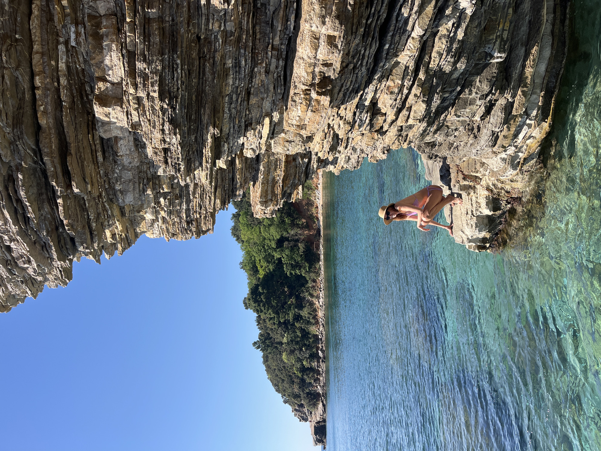A woman in a bikini and hat crouches on rocks at the edge of clear, turquoise water in Budva, Montenegro. The scene is framed by layered rock formations overhead and a tree-lined coastline in the background, under a clear blue sky.