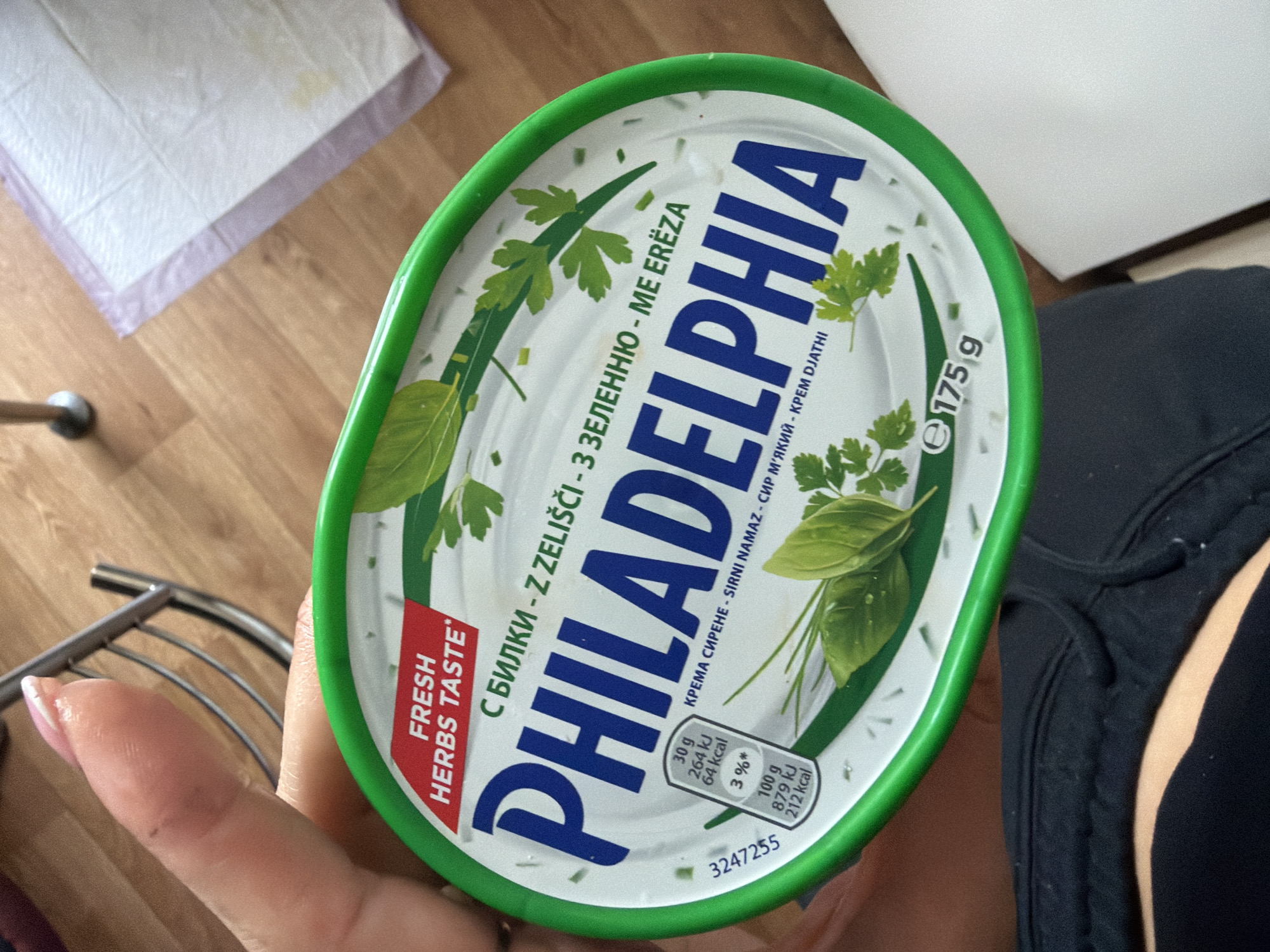 A person is holding a round, green-rimmed container of Philadelphia cream cheese with herbs. The lid is white with green lettering and images of herbs. The product name "PHILADELPHIA" is prominently displayed in large blue letters. Nutritional information and the weight "e 175 g" are also visible. The background is a wooden floor with a purple and white pad. The image does not provide any information to indicate the location is in Kyiv, Ukraine.