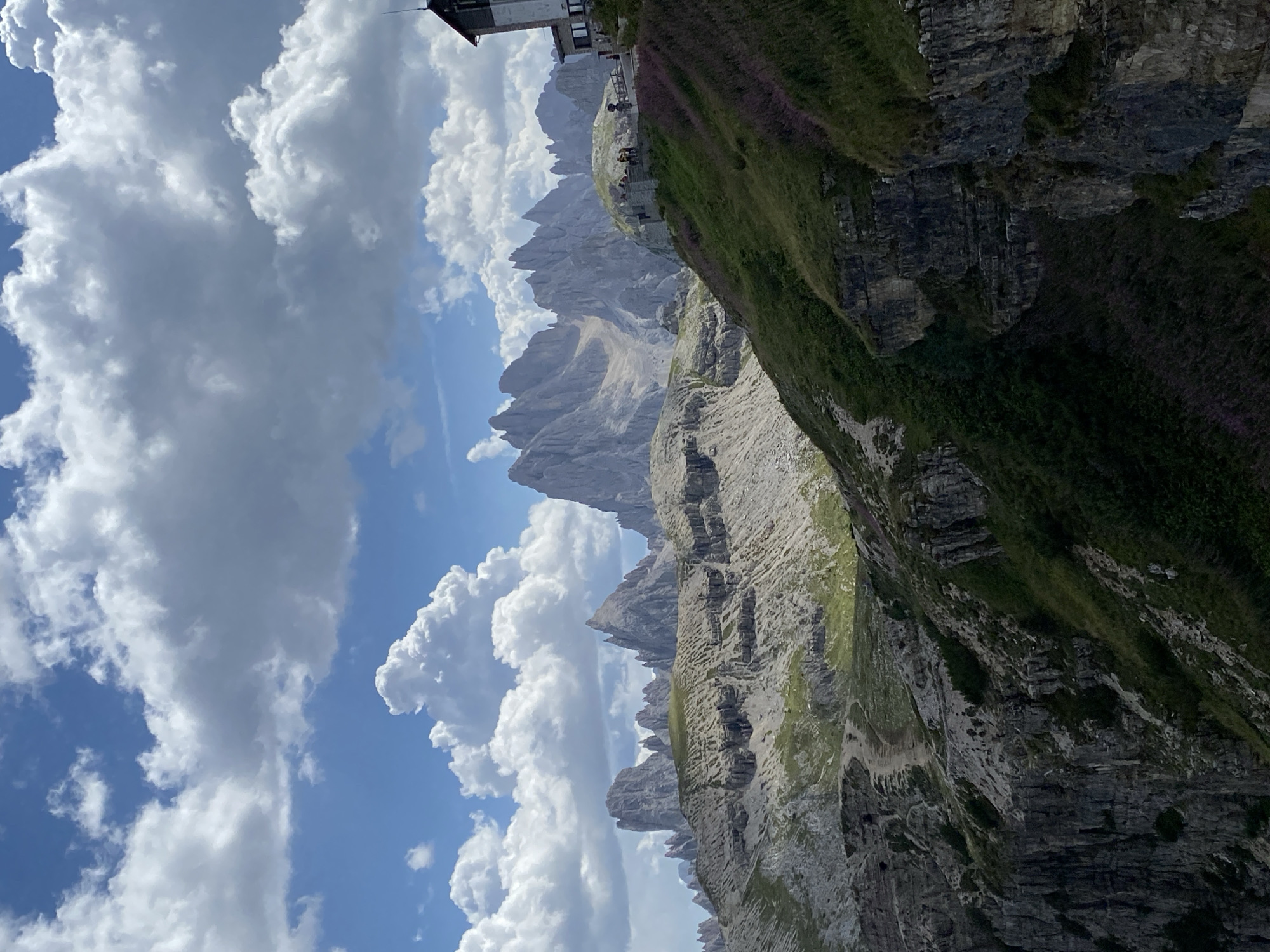 This is a landscape photograph taken in Auronzo di Cadore, Italy, showcasing the dramatic Dolomite mountain range. The image features towering, jagged peaks in the background, bathed in sunlight. In the foreground, a lush green mountain slope is covered with grass and small purple flowers. A stone building, possibly a mountain refuge or lodge, is visible on the right side of the image, perched on the hillside. Several people are gathered near the building on an elevated terrace, looking out at the scenery. A dirt path winds its way up the mountain in the mid-ground. The sky above is a vibrant blue, dotted with fluffy white clouds. The overall scene depicts a scenic mountain vista with elements of human presence.