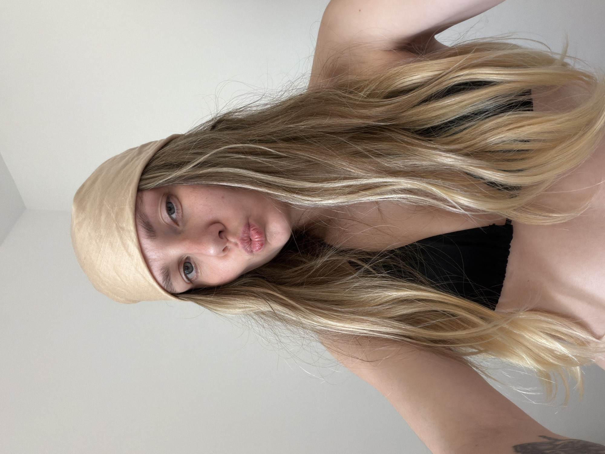 A selfie of a fair-skinned person with long, blonde hair wearing a light brown headscarf and a black top. The person is looking at the camera with a slight pouting expression. The background is a plain white wall. The image does not provide information about the location being Tel-Aviv, Israel.