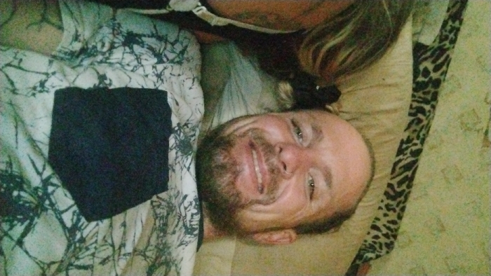 The specific location within the city of Mariposa, United States, is not discernible from the image; it appears to be an indoor residential setting.
The scene features two individuals positioned closely, likely lying down. The primary subject is a male adult with light skin, a full beard, and a ring piercing in his left nostril. He is smiling broadly, revealing teeth, and looking directly towards the camera. He wears a light-colored t-shirt with a dark, abstract, or camouflage-like pattern and a prominent solid black rectangular patch on the chest. To the left of the male subject, a second individual's long, light-colored hair (blonde or light brown) is visible, along with a portion of their left shoulder and a thin white strap, possibly from a bra or tank top. The second individual's face is not visible.
The environment appears to be a bedroom or a comfortable lounge area. Visible objects include a light-colored pillow behind the male subject's head and a distinct black and tan leopard-print fabric, likely a blanket or bedding, in the upper portion of the frame. The lighting is soft and even, consistent with indoor ambient light. No specific actions or interactions beyond their shared proximity and the male's direct gaze at the camera are evident.