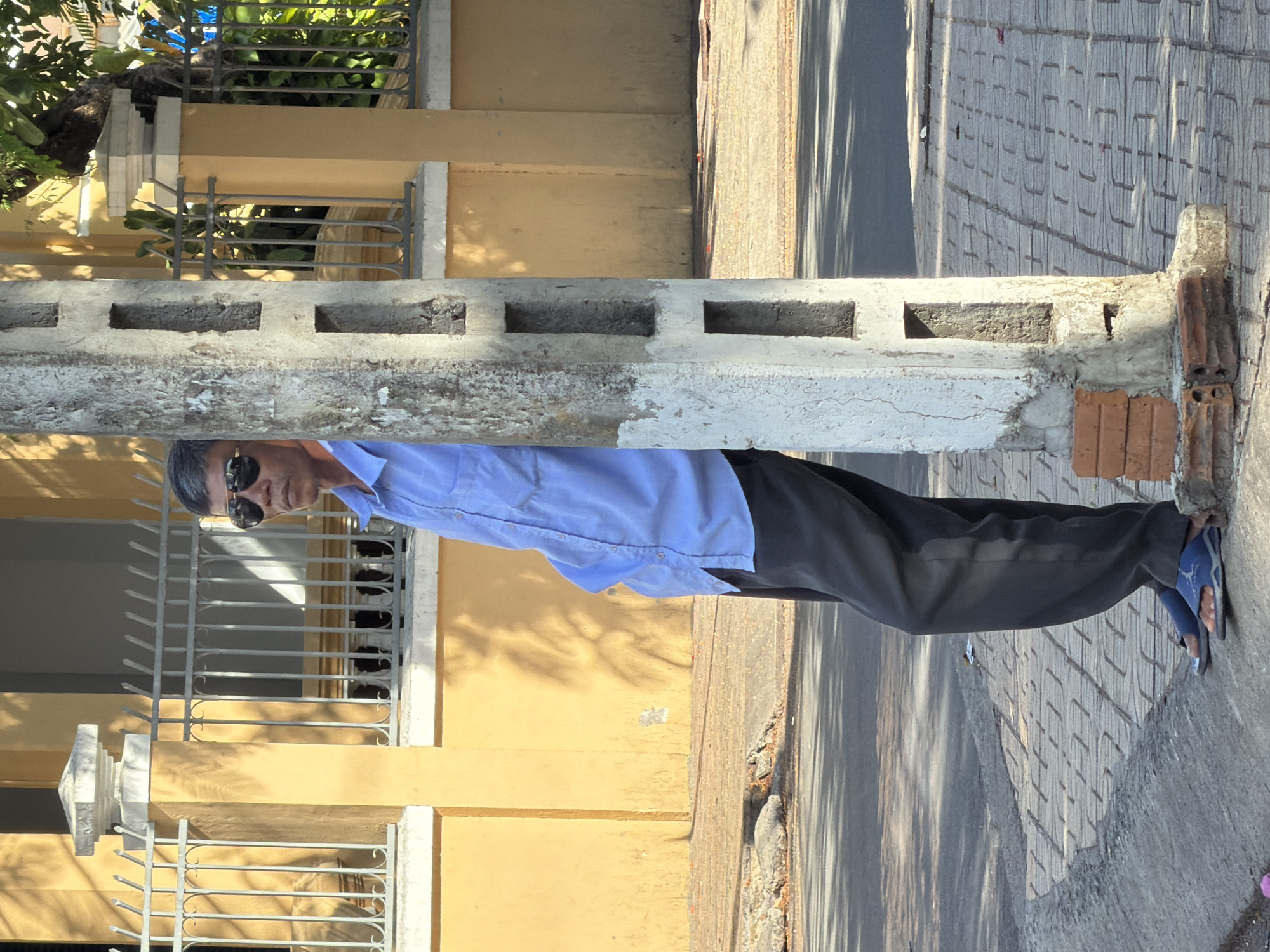 A middle-aged Vietnamese man with graying hair, wearing a light blue button-down shirt and dark gray trousers, stands partially obscured by a concrete utility pole. He is leaning against the pole, with his left hand in his pocket and his head turned slightly to his right, looking downwards. He wears dark sunglasses. The scene appears to be on a sidewalk in Nha Trang, Vietnam, indicated by the man's attire, the style of the building in the background with its yellow facade and ornate metal fencing, and the tropical foliage visible. The ground is paved with patterned tiles, and the utility pole is weathered with visible rectangular openings. A street is visible to the left of the sidewalk.