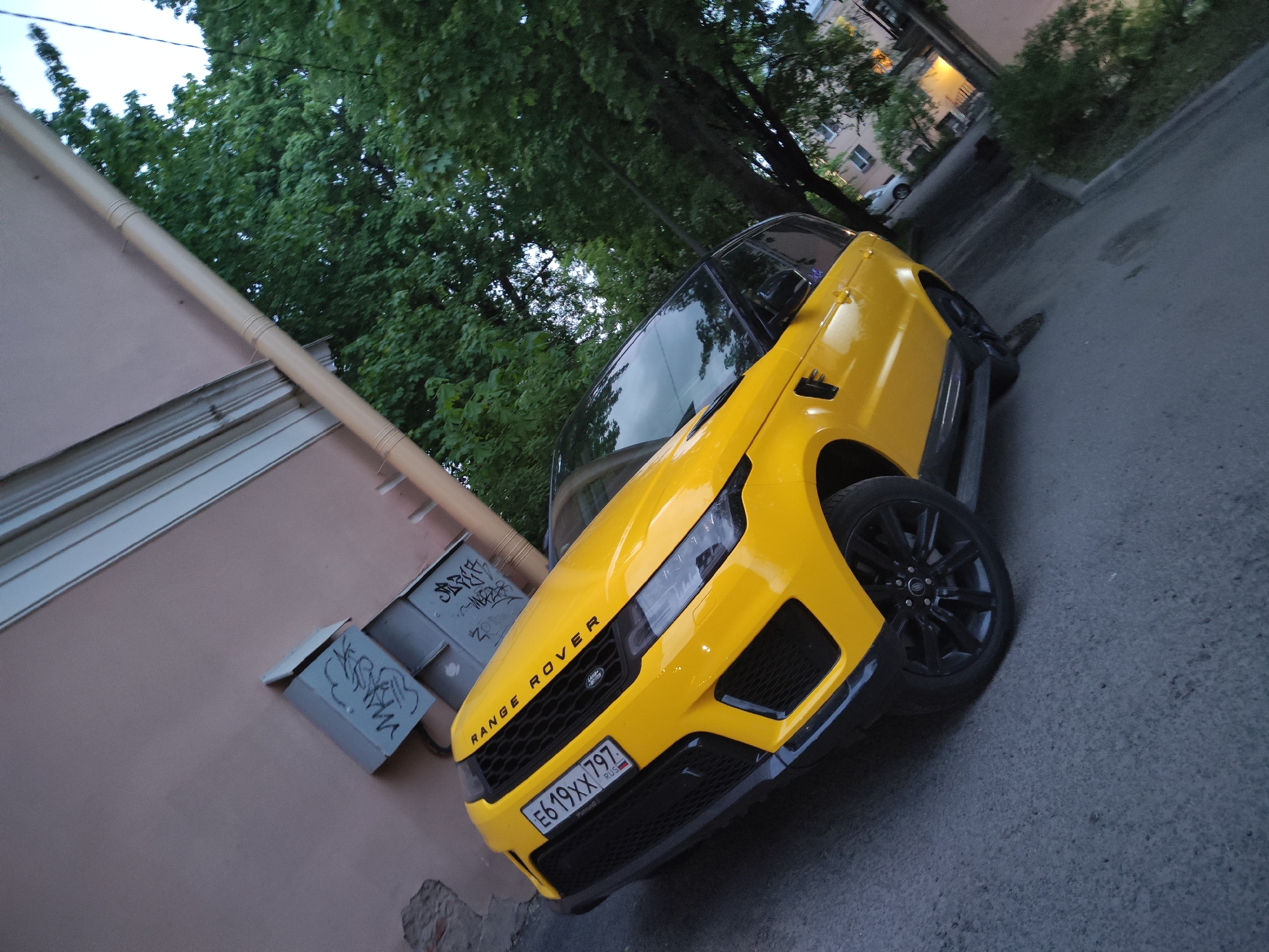 The image depicts a yellow Range Rover parked next to a building in what is reported to be Pushkin, Russia. The car is angled towards the viewer, displaying its front grill, headlights, and Russian license plate number "E619XX797". The vehicle is located on a paved street. Next to the car and connected to a light-colored building, there are two gray utility boxes with graffiti. Above, a tree canopy is visible. The photograph seems to be taken in the evening or during dusk.