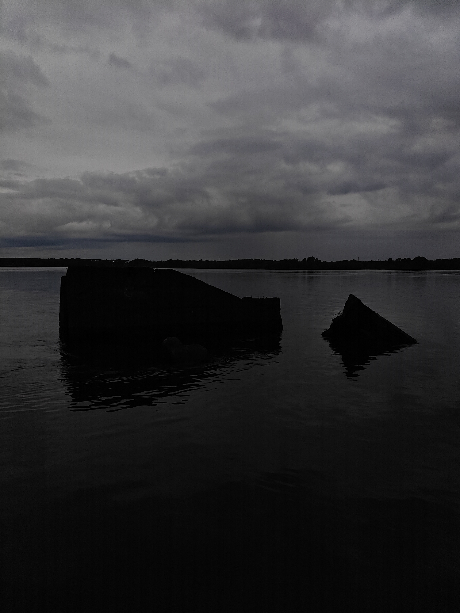 The photograph depicts a gloomy scene at a river in Chaikovsky, Russia, under a gray, cloudy sky. In the foreground, two large, dark, partially submerged concrete structures are visible in the water. The first is more substantial and appears to be a collapsed section, while the second is smaller and triangular. The water surface is dark and slightly rippled. In the background, the opposite shore is lined with trees and vegetation, obscured by the distance and low light. The overall atmosphere is somber and still.