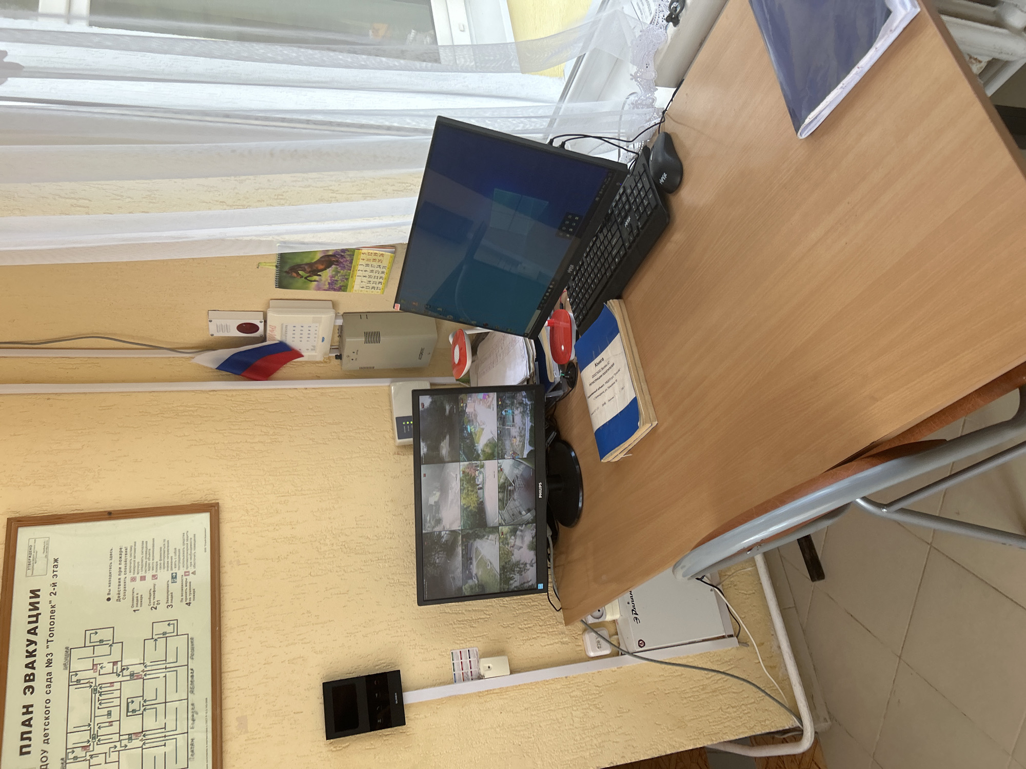 The image shows a desk with computer equipment, a monitor displaying security camera feeds, and various documents. A fire evacuation plan for a kindergarten in Gelendzhik, Russia, is prominently displayed on the wall. The desk is made of wood and appears to be in an office or administrative setting within the kindergarten. On the desk, a laptop, keyboard, and mouse are arranged, along with a book and what seems to be a folder. The monitor is displaying multiple camera views, suggesting a security monitoring setup. Several electronic devices, possibly intercoms or alarms, are mounted on the wall. A small Russian flag is also visible, attached to the wall near a calendar. The overall scene suggests an operational environment within an educational institution, likely focusing on safety and security. The lighting suggests it is daytime.