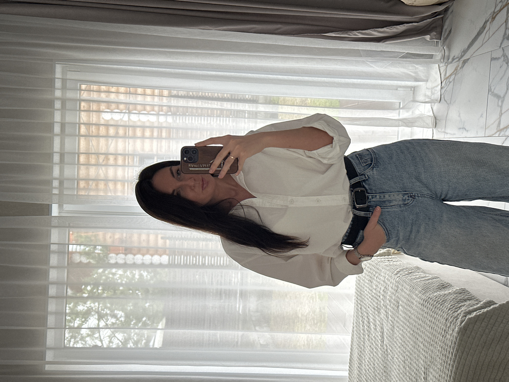 A woman is taking a selfie with her phone. She is wearing a white, button-down shirt and blue jeans. She also has a black belt and a watch on her wrist. The background shows a window with sheer curtains and blinds. Outside the window, trees and buildings can be seen. The setting is a room with a patterned floor and furniture covered with light-colored fabric. The phone case reads "RN40TN993T". While it is impossible to confirm the exact location without further information, elements like building style and foliage may indicate a possible location in Chaikovsky, Russia.