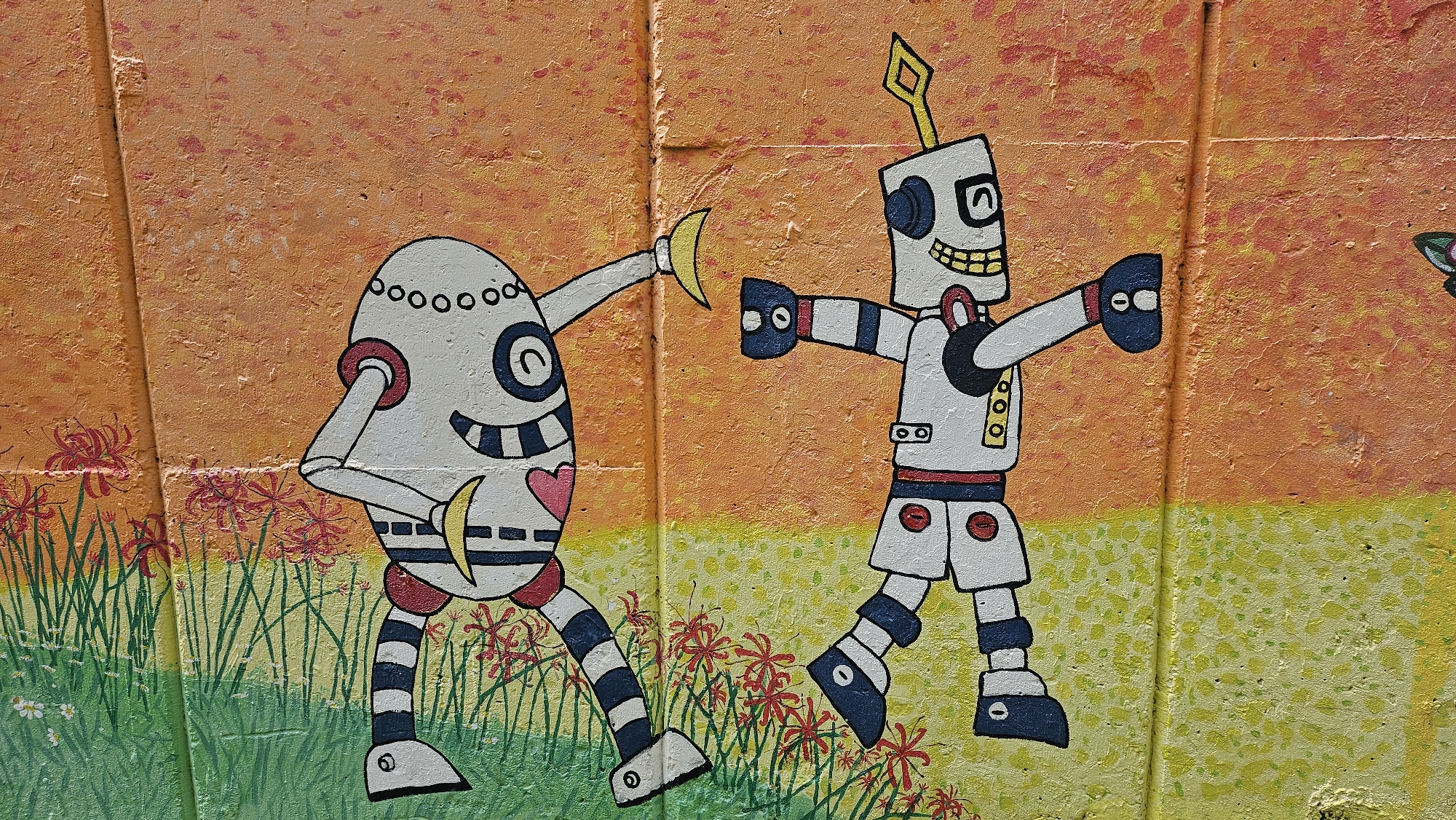 A mural depicts two robots against an orange and yellow background on a textured wall. The robot on the left has an egg-shaped body, black and white striped legs, and a red heart on its chest. It holds a yellow crescent shape, likely a banana, in its right hand. The robot on the right has a rectangular body, a square head with an antenna, and wears shorts. Both robots are smiling. The background includes red spider lilies and green grass painted at the bottom of the wall. The mural is likely located in Ulsan, South Korea.