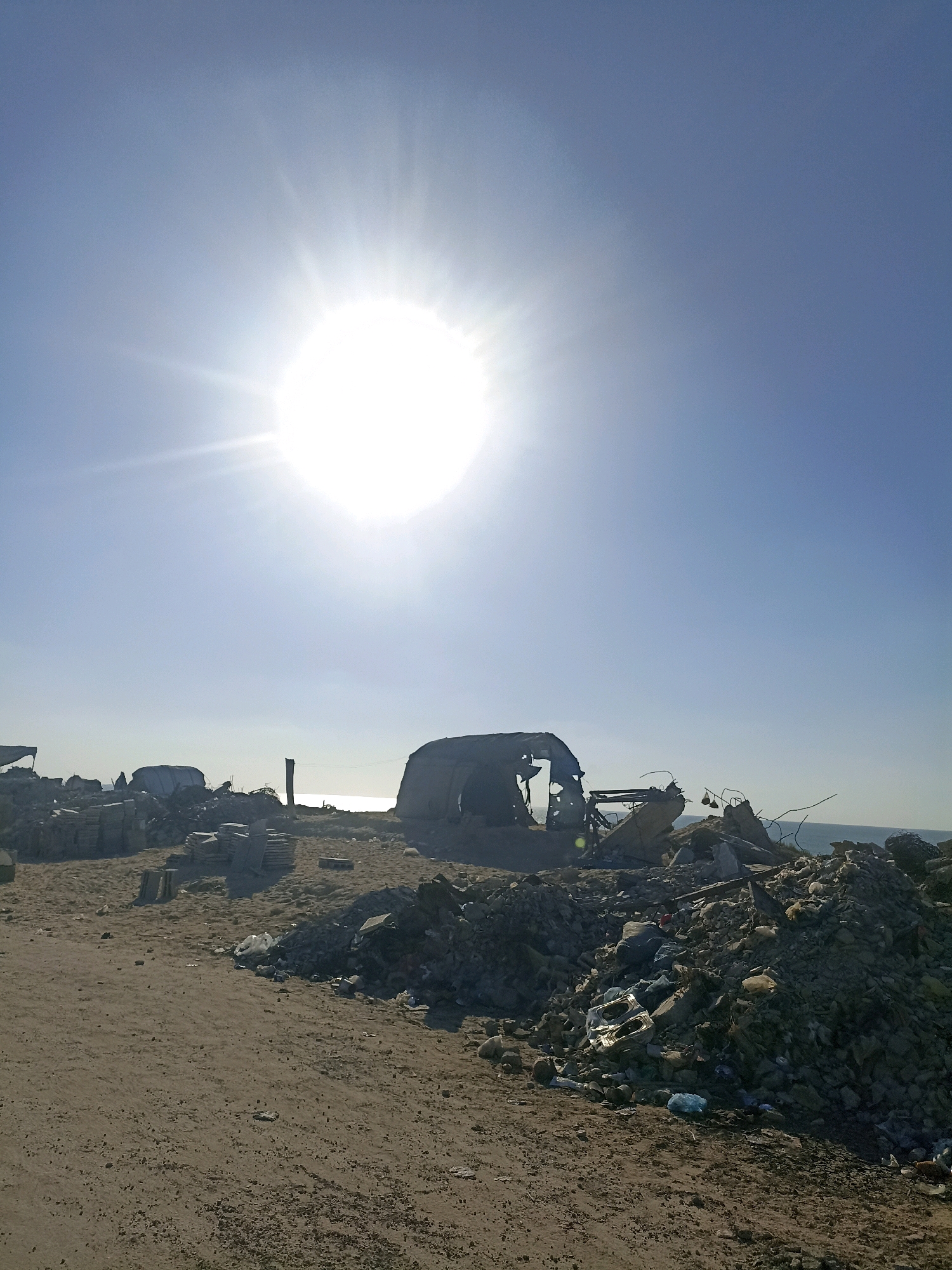A wide-angle, eye-level shot captures a desolate, debris-strewn landscape under a bright, midday sun in Gaza. The foreground features a dirt path leading towards a mound of rubble, intermixed with white plastic bags and other refuse. To the left, stacks of grey building materials, possibly tiles or slabs, are arranged neatly, contrasting with the surrounding chaos.
Dominating the mid-ground are the skeletal remains of what appears to be a vehicle, heavily damaged and broken, with exposed metal framing and a tattered covering. Further back, other structures, some partially collapsed and resembling tents or makeshift shelters, are visible amidst the debris. A solitary, dark vertical pole stands amidst the ruins.
The background reveals the calm, shimmering surface of the sea under a clear, pale blue sky. The sun, a brilliant white orb with visible rays, is positioned high in the sky, casting a bright glare that slightly washes out the upper portion of the image. The overall impression is one of destruction and neglect, with the natural beauty of the sea and sky providing a stark juxtaposition to the man-made devastation on land.