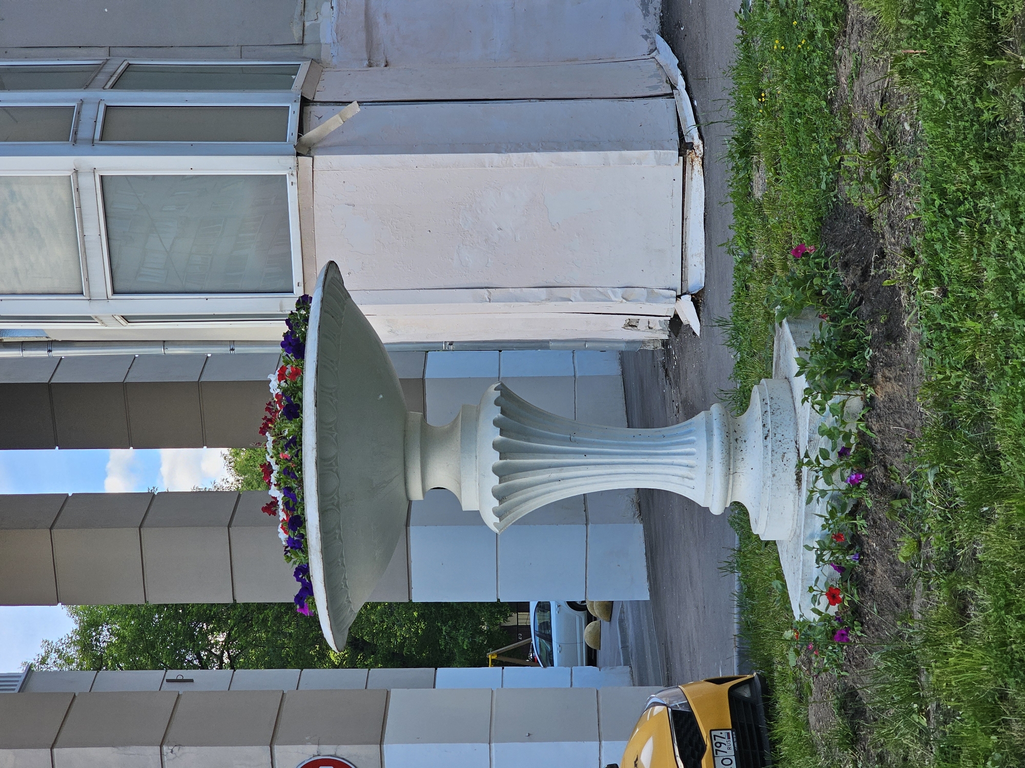 A vertical outdoor shot in Khimki, Russia, captures a large white ornamental flower pot filled with colorful flowers. The pot sits near a building with gray and white rectangular pillars and multi-paned windows. A yellow vehicle, possibly a taxi, is partially visible at the bottom of the frame, with a license plate ending in "797 RUS" is displayed. Grass and greenery line the edge of the scene, and a glimpse of blue sky is visible through the pillars.