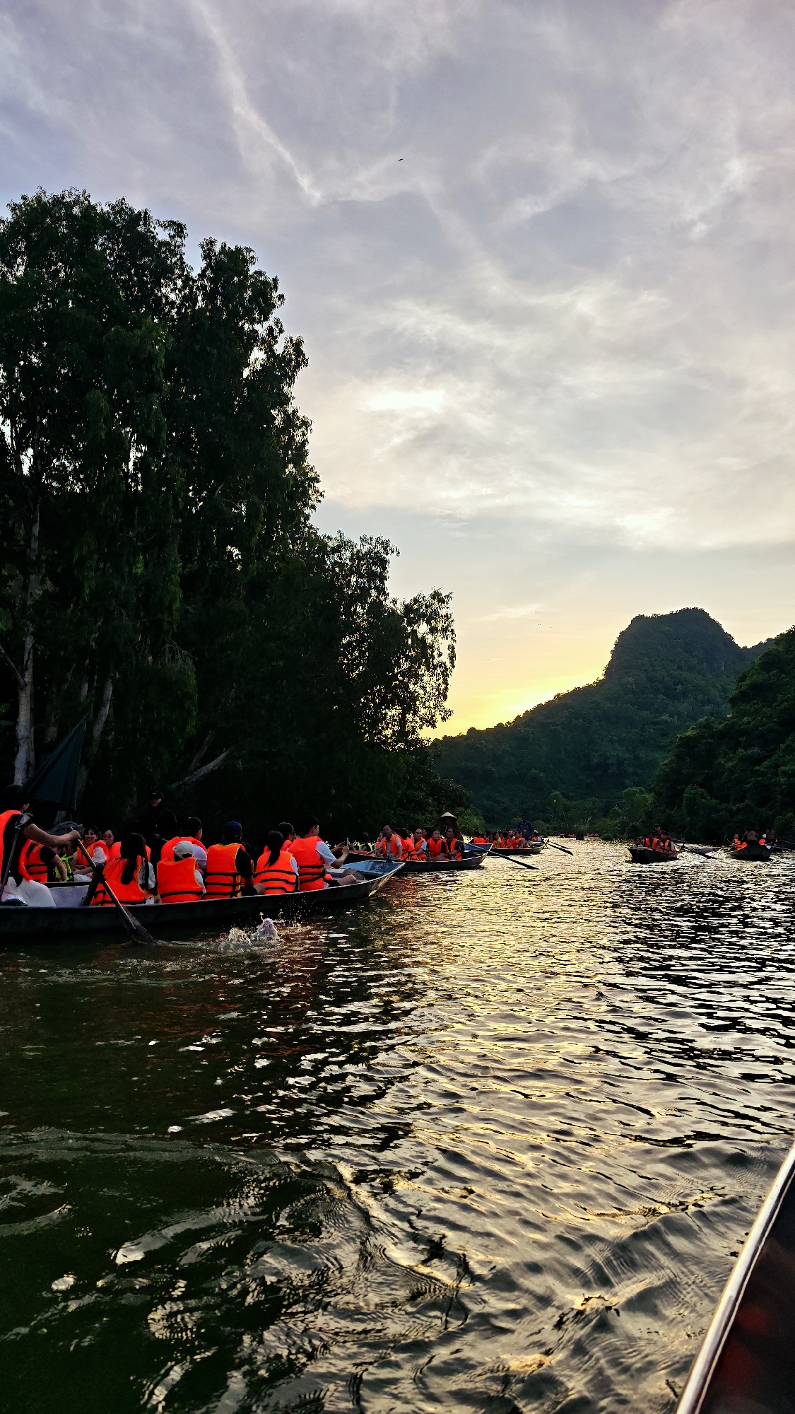 A group of tourists wearing orange life vests are in several boats on a river at sunset, near Yên Sơn Ward, Vietnam. The boats are moving from left to right in the frame. Lush green trees line the left bank of the river, and green, tree-covered mountains are visible in the distance on the right. The sky is cloudy with a warm glow from the setting sun. The water reflects the light and the surrounding landscape, creating rippling patterns.