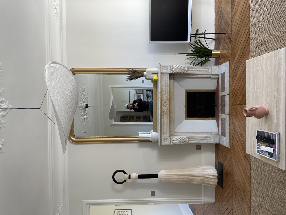 The image depicts the interior of a Parisian apartment, characterized by its classic Haussmannian architectural elements. The central focus is a white marble fireplace with ornate carvings and a dark, empty firebox, above which a large, gold-framed mirror is mounted. A white, irregularly shaped modern pendant light fixture hangs from the high ceiling, which features intricate white plaster moldings. The pendant light is reflected in the mirror.
In the reflection, a person is visible, holding a smartphone to take the photograph. The individual wears a dark top and a light-colored cap.
To the left of the fireplace stands a tall, floor-standing sculpture comprising a black ring at the top, alternating light wooden and black cylindrical sections, and a large, light-colored fiber tassel-like base resting on a dark square platform. A white vase is placed on the left side of the fireplace mantel. A doorway on the left leads to another room with white walls and a framed artwork visible in the distance.
On the right side of the fireplace mantel, a uniquely shaped white vase holds dried lavender sprigs, accompanied by a single yellow lemon. Further to the right, a modern flat-screen television with a white bezel stands on a black tripod mount. Adjacent to the television, a snake plant in a golden pot rests on the floor.
In the foreground, a light beige travertine coffee table sits on a light brown woven rug. On the table, a book or magazine displays a remote control and another small dark object. A small, reddish-orange ceramic pitcher or vase is also on the table. The flooring throughout the visible space consists of polished light brown herringbone parquet. The walls are uniformly white.