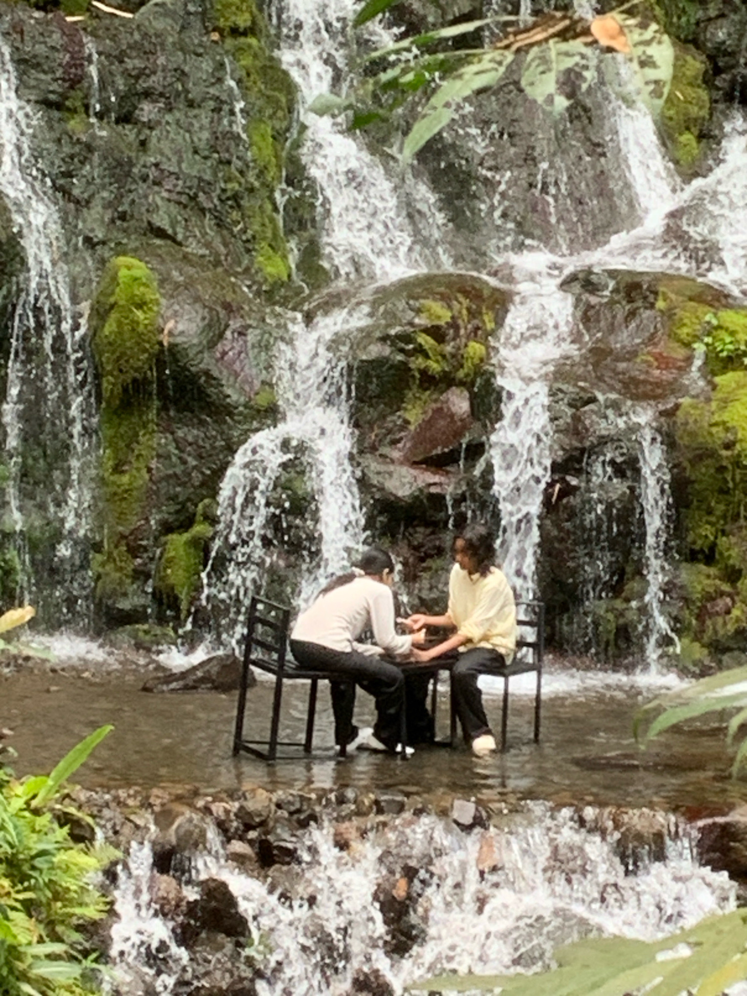 Two individuals are seated at a table in front of a waterfall, likely at the Pronojiwo Waterfall in Pronojiwo, Indonesia. The waterfall cascades down a rocky, moss-covered cliff face. The individuals are positioned such that the water flows around their legs and the base of the table. The person on the left wears a light-colored long-sleeved shirt and dark pants, while the person on the right is dressed in a yellow long-sleeved shirt and dark pants. They are both seated on chairs and appear to be engaged in a conversation or activity at the table. The foreground of the image shows the shallow water flowing over rocks, with greenery present on the left and right sides, framing the scene.