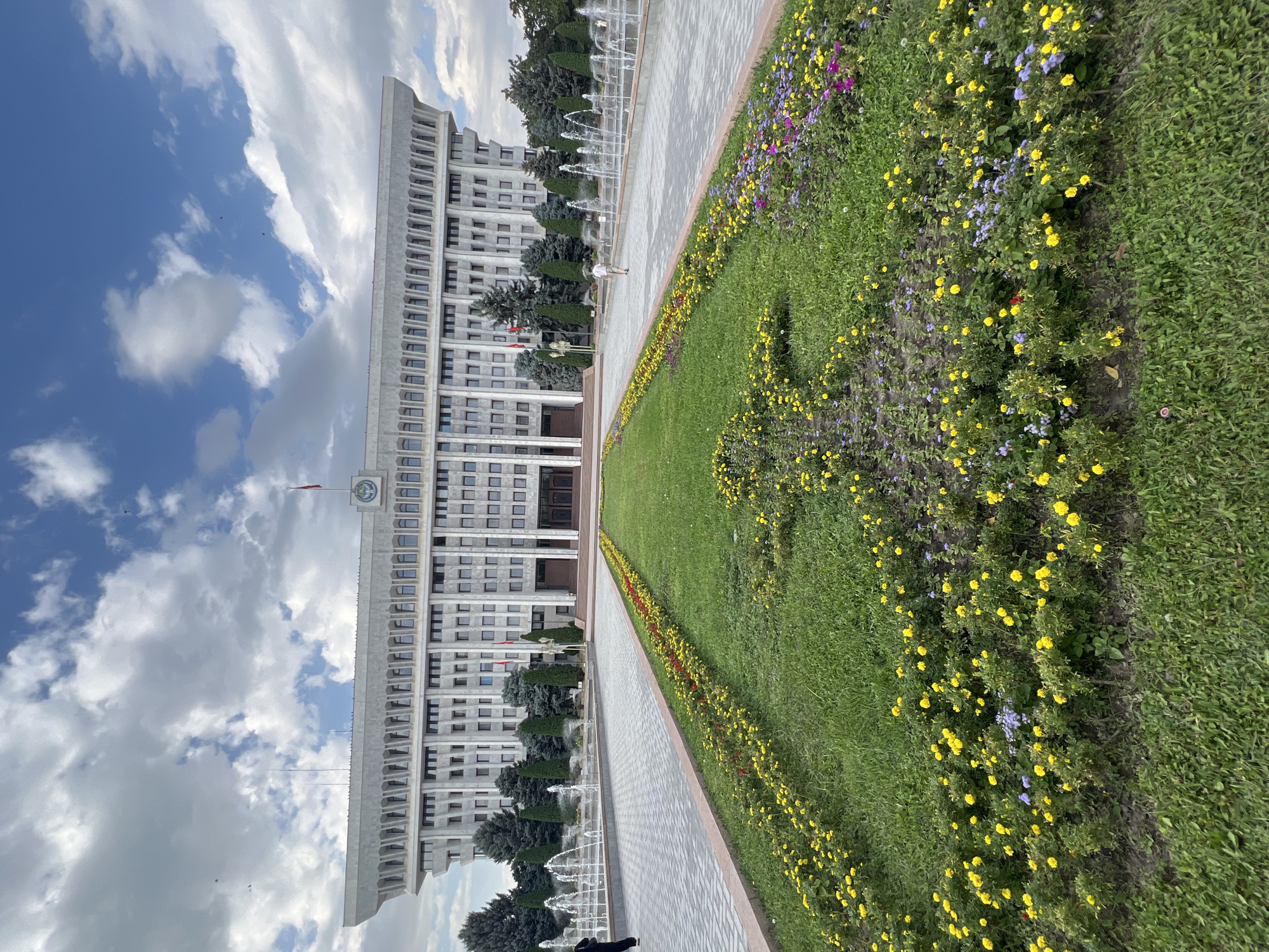A wide-angle, low-angle exterior shot captures the Ala-Too Square in Bishkek City, Kyrgyzstan, under a cloudy sky. The focal point is the White House, a large, imposing government building with a distinct angular facade and numerous windows. In front of the building, a symmetrical layout features two long pathways bordered by a central strip of meticulously maintained green grass and vibrant flowerbeds. The flowerbeds are densely planted with a mix of yellow and purple flowers, with some red accents.
Fountains are actively spraying water along the pathways on either side, creating a lively atmosphere. A few individuals are visible in the scene. On the left pathway, a person in black pants is walking away from the camera. On the right pathway, a young person in a white t-shirt and shorts is walking towards the building. The architecture of the White House is characteristic of Soviet-era modernist design, with its concrete structure and repetitive geometric patterns. The overall impression is one of grandeur and order, with the natural beauty of the flowers and the activity of the fountains adding a touch of vibrancy to the formal setting.