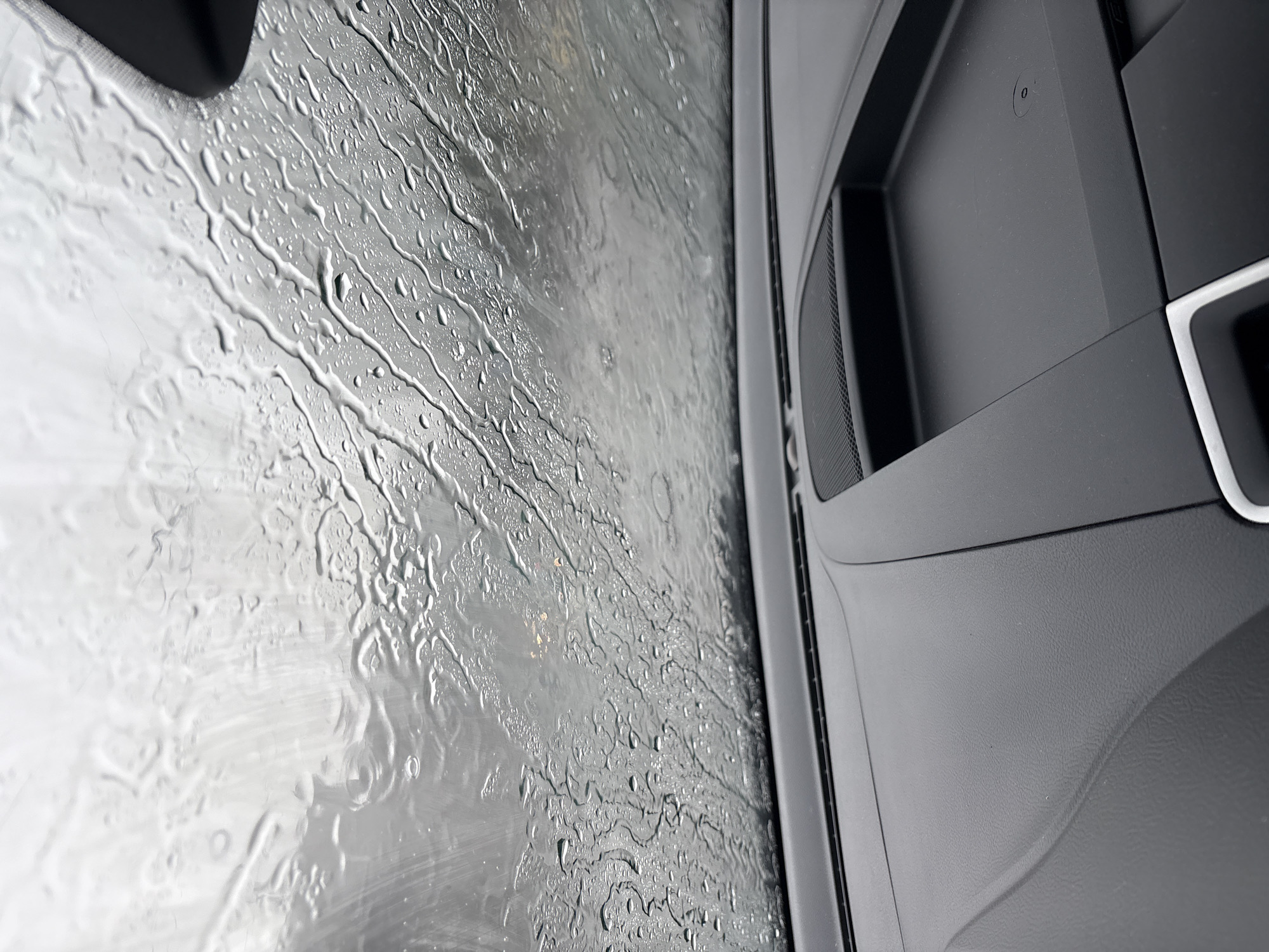An interior shot of a car looking out through a rain-streaked windshield. The dashboard, composed of dark textured plastic, is visible in the foreground. A speaker grille is integrated into the dashboard on the left side, and a shallow storage compartment with a circular indentation is present on the right. The windshield is heavily distorted by numerous raindrops and rivulets of water, creating a blurred and abstract view of the overcast sky and distant, indistinct urban lights, possibly in Vitsebsk, Belarus.