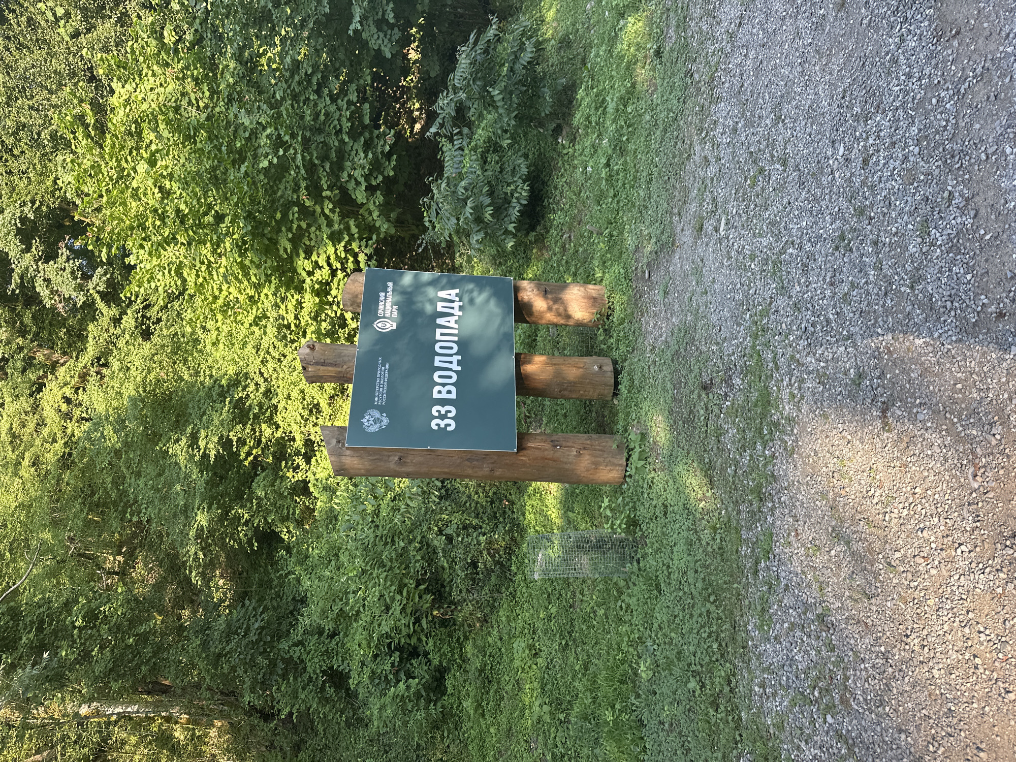 The image depicts a sign in a natural, wooded environment. The sign, which is a rectangular green board mounted on three wooden posts, displays text in Russian. The text includes "33 ВОДОПАДА" (33 Waterfalls), and smaller text at the top which appears to be official emblems or names of organizations. The sign is situated in an area with dense green foliage and trees in the background, and a gravel path in the foreground. There is a wire mesh cage protecting a small plant to the left of the sign. The overall scene suggests a natural park or a tourist attraction. The location is identified as the city of Bol'shoi Kichmai, Russia, likely indicating a specific natural landmark or trail within that area. There are no people or animals visible in the image.