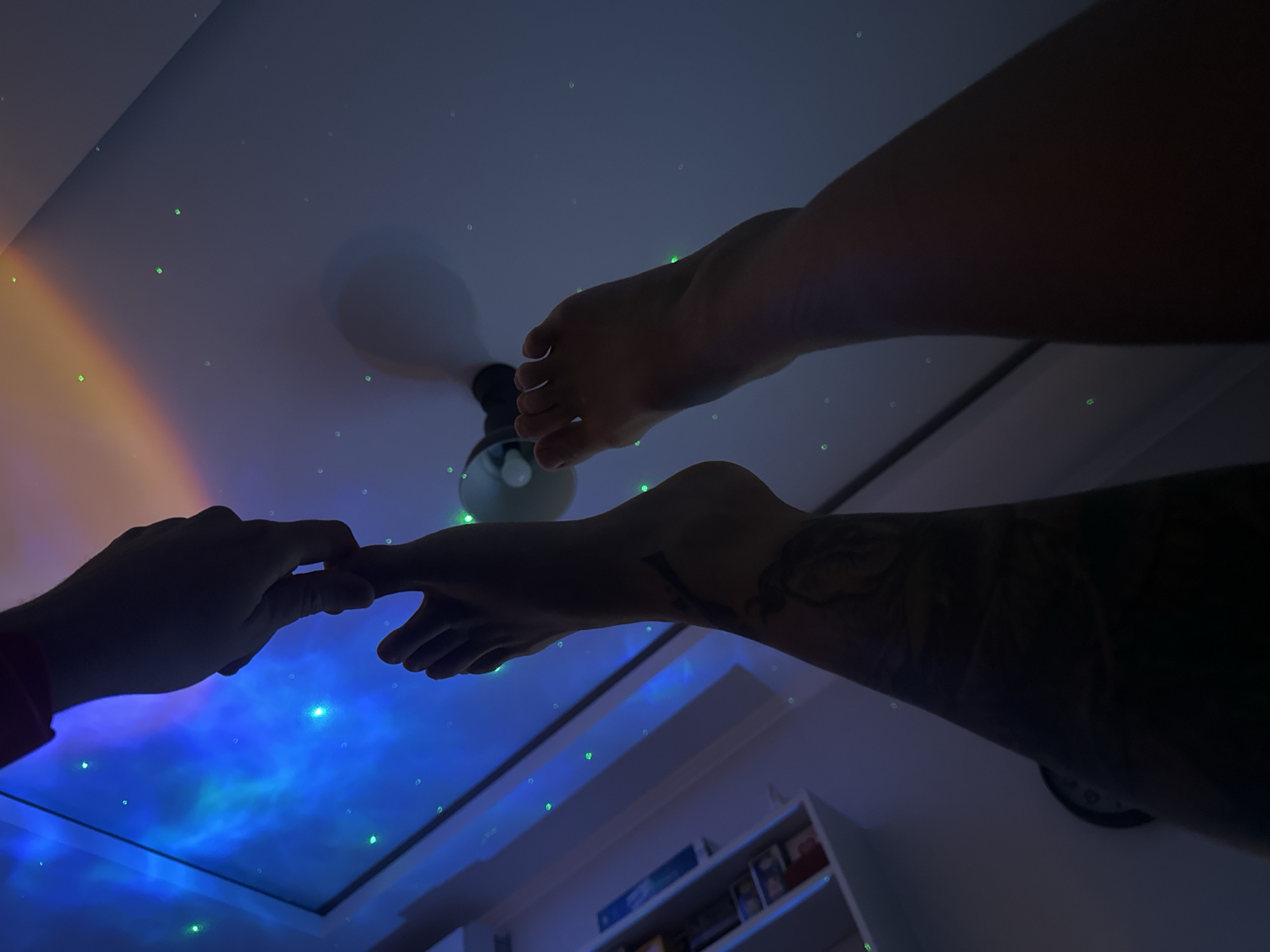 The image shows a low-angle perspective of two people's feet and hands interacting in a room illuminated by a galaxy projector. The projector casts blue and green light patterns resembling a nebula and stars onto the ceiling and walls, with a warm orange and yellow gradient visible in the upper right corner. One person's bare feet are visible, with tattoos on the leg of the person whose feet are closer to the bottom of the frame. The other person's hand is reaching down to touch the toes of the first person. A ceiling fan is partially visible in the background, and shelves with books and objects can be seen on the left side of the frame. The scene appears to be an indoor personal space, likely a bedroom or living area, designed for ambiance. There is no indication of the specific location being in Moscow, Russia.
