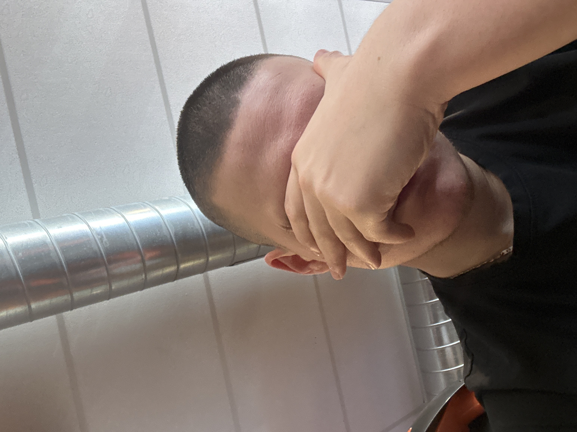 A single adult male with a short buzz cut haircut is depicted in an unidentified indoor location within Schagen, Netherlands. The subject occupies the majority of the frame, from the chest up, wearing a black short-sleeved t-shirt and a gold-colored chain around the neck. The subject's right hand is positioned over the face, specifically covering the eyes and upper nasal area. The background features a large, cylindrical, spiral-seamed metallic ventilation duct running diagonally from the upper left to the lower right. The ceiling or upper wall is a light, uniform color, exhibiting subtle horizontal panel lines. Overhead artificial lighting is evident. A segment of another horizontal metallic fixture is visible behind the subject's left shoulder (viewer's right). The subject's left ear is partially visible.
