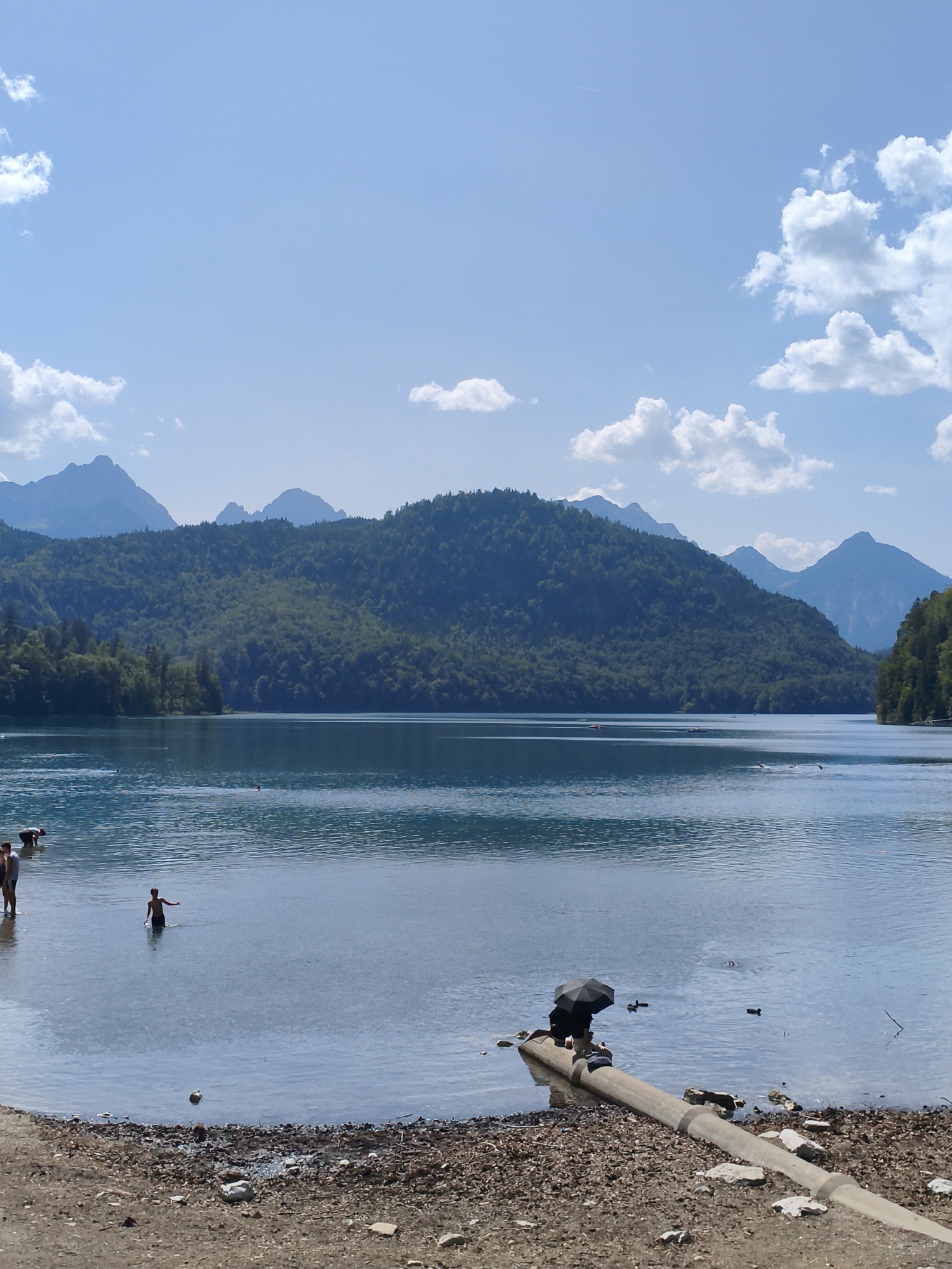 A scenic view of a lake in Schwangau, Germany, on a bright, sunny day with a clear blue sky and scattered white clouds. The foreground is a rocky, pebbly shore leading to the calm, blue water. Several people are visible in the lake, some wading and others swimming. In the middle ground, a person sits on a large concrete pipe that extends into the water, holding a black umbrella. In the background, a densely forested mountain range rises majestically, with several prominent peaks silhouetted against the sky. The water is tranquil, reflecting the sky and the surrounding landscape. The overall impression is one of a peaceful and idyllic summer day by the lake.