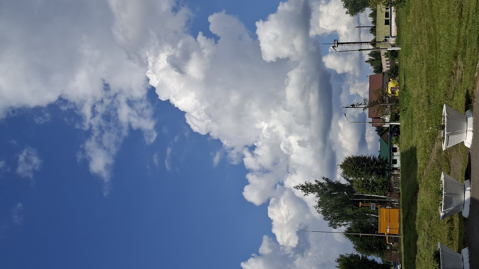 This image depicts a bright, sunny day in Myadzyel, Belarus, with a vibrant blue sky filled with large, fluffy white clouds. In the foreground, a grassy area is visible, with three white planters containing sparse green foliage. Beyond the grass, a paved area with a few parked cars is present. Several buildings are scattered across the mid-ground, including a prominent yellow structure with an orange fence, likely part of industrial infrastructure, and other buildings with green and brown roofs. Trees of varying heights and types are interspersed throughout the landscape. Tall poles with what appear to be antennas or communication equipment are also visible. The overall scene suggests an outdoor environment on the outskirts of a town or in a semi-industrial area.