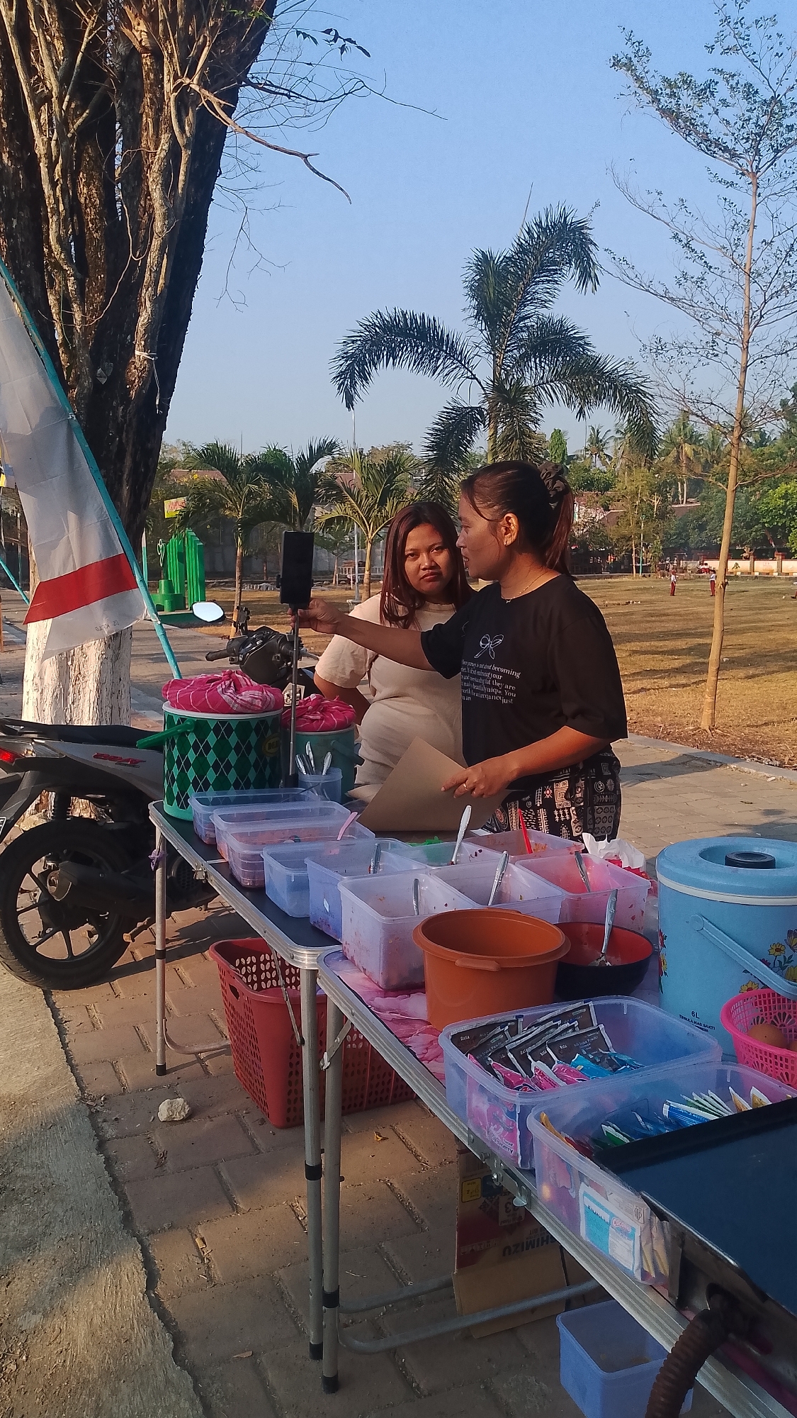In Karanglangu, Indonesia, two women are standing behind a foldable table laden with various food items and containers. One woman, wearing a black t-shirt with a graphic design and patterned pants, holds a smartphone on a selfie stick and appears to be speaking to the other woman. The second woman, dressed in a light-colored t-shirt, looks towards the selfie stick. The table displays plastic containers filled with what appear to be food ingredients, possibly for snacks or drinks, along with bowls and utensils. To the left of the table, a motorcycle is parked, and a white flag with red and white stripes is visible. In the background, palm trees and other vegetation suggest a tropical or subtropical environment. The ground is paved with interlocking stones. The overall scene depicts a vendor stall, possibly at an outdoor market or event.