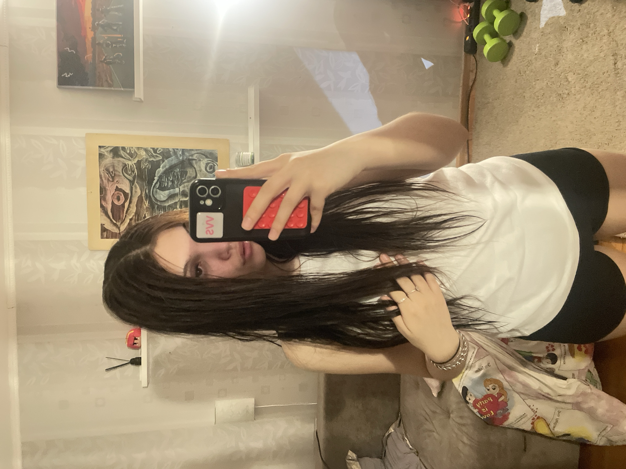 A young woman is captured in a mirror selfie within an indoor setting in Saint Petersburg, Russia. She has long, dark hair, a neutral expression, and is wearing a white t-shirt and black shorts. Her left hand holds a black smartphone, featuring a distinct red, brick-like attachment and a white sticker on the back that reads "2vv" (as reflected). She also wears rings on her right hand and a silver-colored bracelet on her left wrist.
The reflection reveals details of the room. The background wall is light-colored with a subtle floral or leaf pattern. Two framed artworks are visible: an abstract painting with a prominent, eye-like central feature in grey and pink tones, and above it, a landscape depicting a sunset or sunrise with birds, rendered in warm and dark hues. In the upper right of the reflection, three neon green dumbbells are stacked, alongside a string of red fairy lights. A bright glare from an artificial light source is visible near the top center. The floor appears to be a light brown, textured carpet or mat. A grey piece of furniture, likely a couch or bed, is partially covered by a colorful blanket adorned with cartoon characters, including what appear to be Disney princesses, and the visible text "PARTY IS LOVE". On the left side of the reflection, a white roll on a holder is visible against the patterned wall.