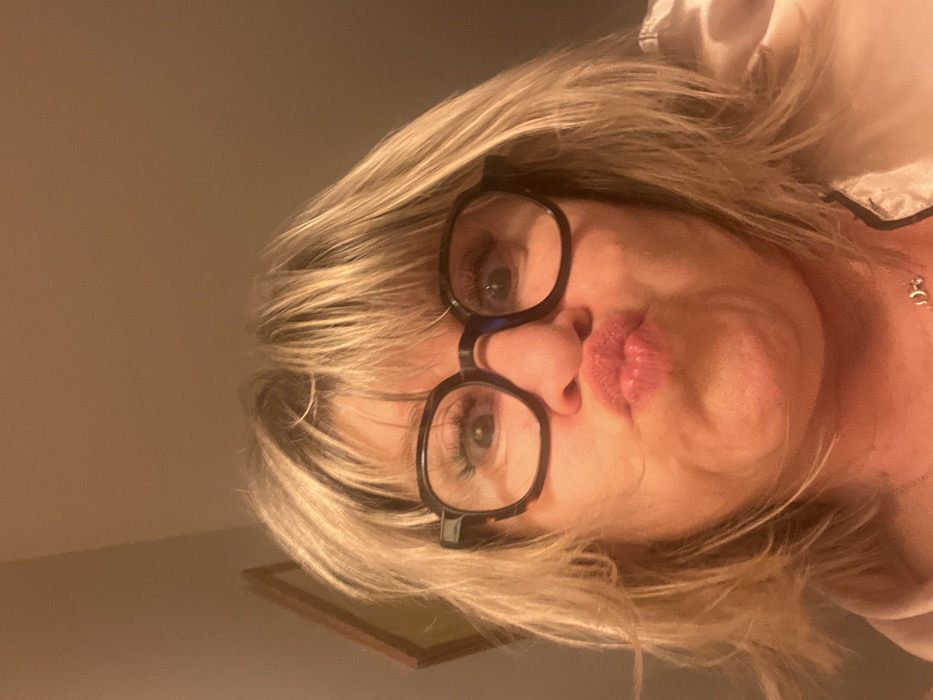 A woman with blonde hair and glasses puckers her lips at the camera. She is wearing a light-colored top or robe, and a delicate necklace is visible. The photo is taken from a low angle, looking up at her face. A framed picture is partially visible in the background. The lighting appears warm and soft.