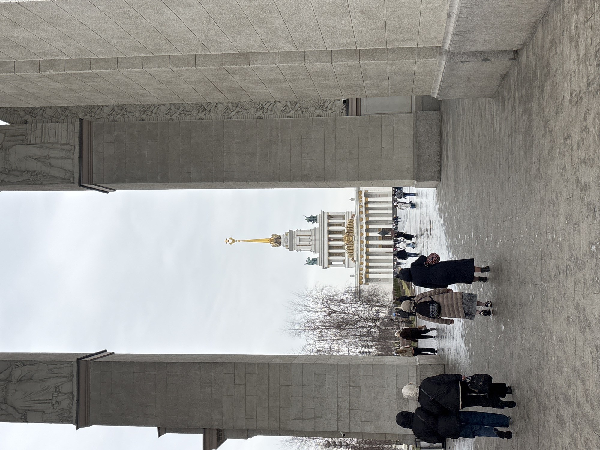 The image shows a view through a concrete archway towards a large building topped with a golden spire, likely the Main Pavilion at the All-Russia Exhibition Centre (VDNKh) in Moscow, Russia. People are walking on the pavement towards the building. The sky is overcast, and the foreground is composed of the archway's textured concrete surfaces. The building has multiple tiers of columns and statues. Bare trees are visible in front of the building.