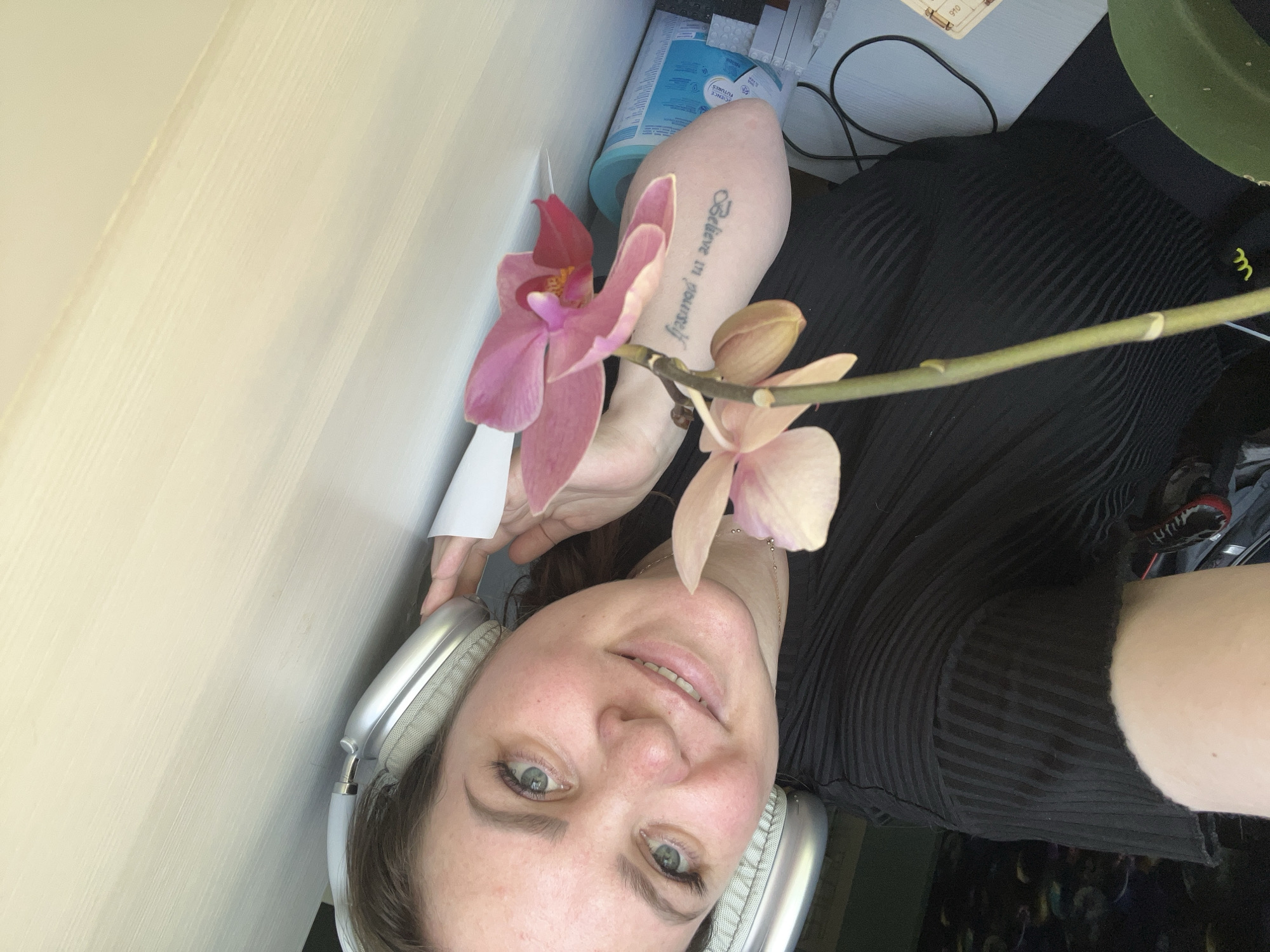 A woman with fair skin and green eyes smiles into the camera while wearing silver headphones. She is holding a light pink orchid with multiple blooms and an unopened bud. The orchid is near her left arm, which has a tattoo that reads "Believe in Yourself". She is wearing a black, vertically striped top. In the background are a white desk, a blue and white container, some papers, and a black cable. The image is taken indoors. There is no indication that the location is Noviy, Russia.