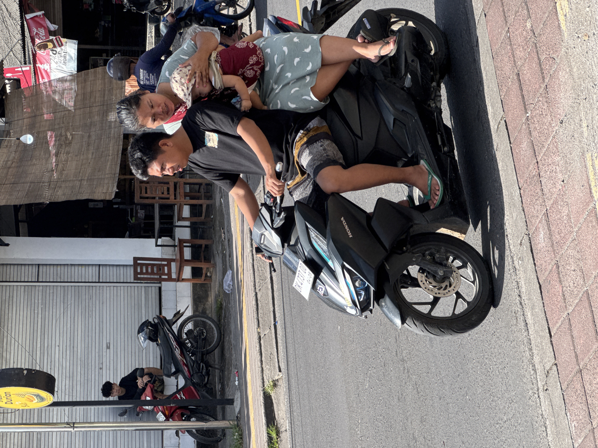 The image captures a scene on a street in Kuta Selatan, Indonesia, featuring a family of three on a black Honda motorcycle. The father is seated on the motorcycle, wearing a black shirt and shorts. The mother sits behind him, holding a young child wearing a patterned shirt. Both parents are not wearing any visible safety equipment.
The motorcycle is parked on the side of a paved road with a brick-paved sidewalk to the right. The motorcycle's license plate is partially visible, reading "DK 4737 DD" and showing the date "04-28". In the background, there is a building with a partially closed shutter door. Additionally, another parked motorcycle can be seen in the background near a street sign.