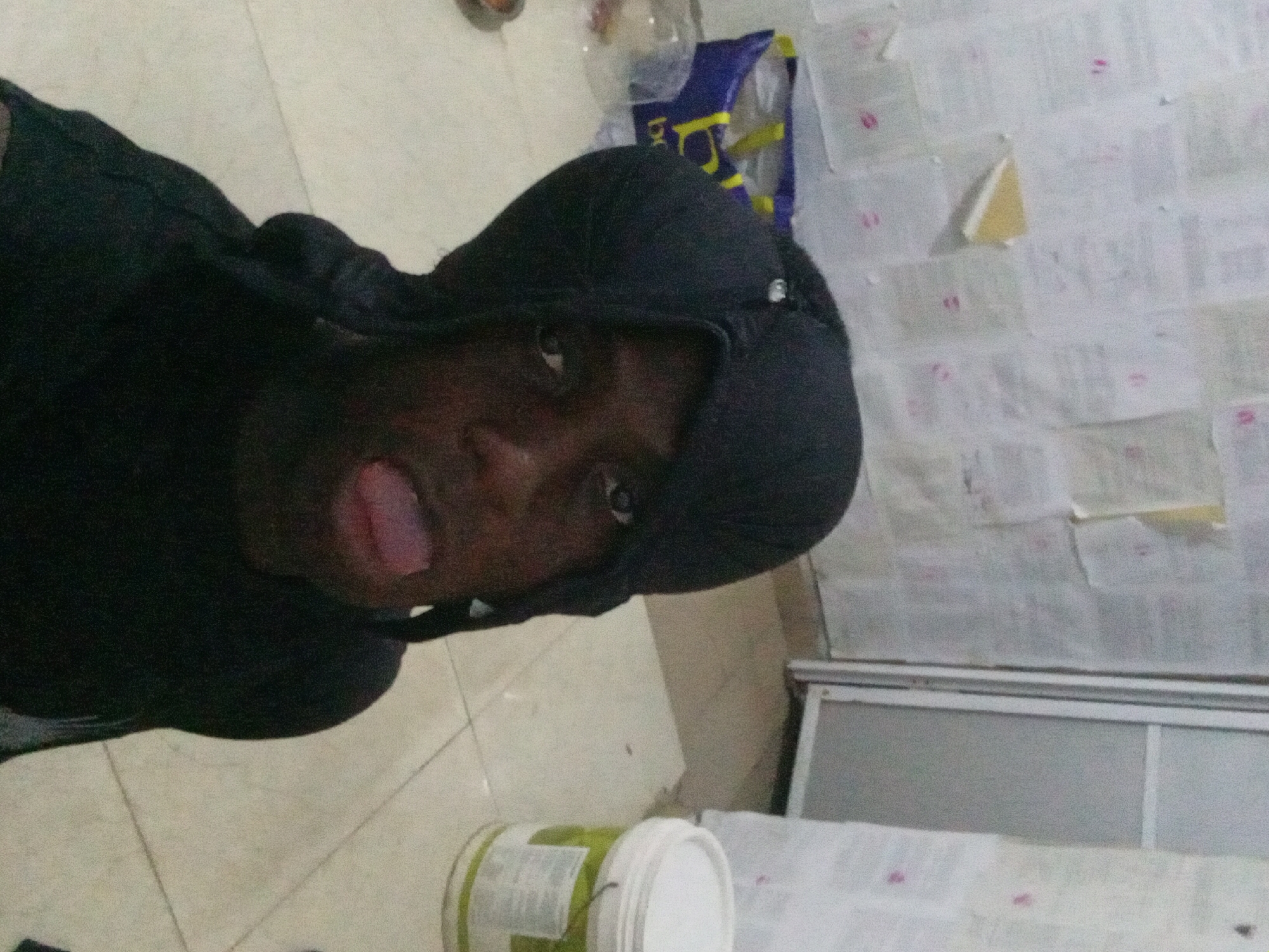 A person with dark skin and a black hooded sweatshirt is sticking their tongue out at the camera. They are lying on the floor in a room with beige tiles. Behind the person is a white wall covered in what appears to be small pieces of paper. There are also several containers and bags near the wall. The location is most likely Akwete, Nigeria.