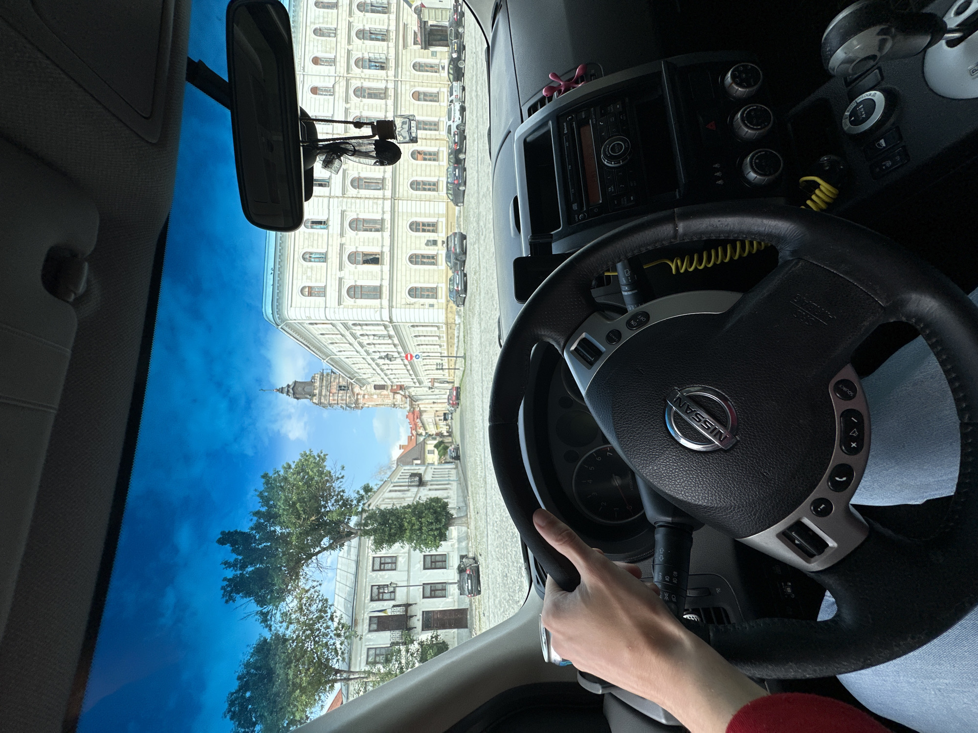 The image is a first-person view from the driver's seat of a car in Kamianets-Podilskyi, Ukraine. The driver's hands are on the steering wheel, which has a Nissan logo. The dashboard and car interior are visible. Through the windshield, a city square is seen with several cars parked. In the background, there are historic buildings with arched windows and a prominent church tower. Trees line the square, and the sky is blue with some clouds. The scene suggests a sunny day in a European city.