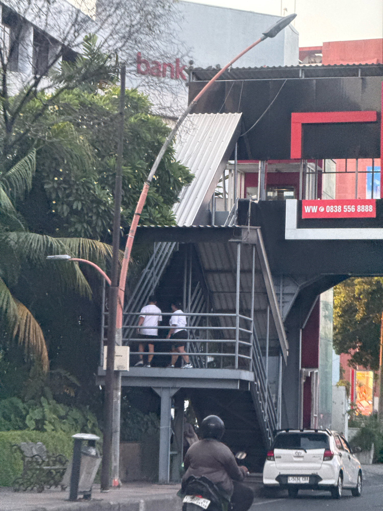 This image depicts an outdoor urban scene in Surabaya, Indonesia. In the foreground, a person on a motorcycle is visible from behind, wearing a helmet. To the right, a white Toyota car is parked on the street, with its license plate partially obscured. Behind the motorcycle and car, a multi-level pedestrian overpass is prominent. The overpass has a metal structure with stairs and railings, and two individuals in white shirts are standing on it. Above the overpass, a building with a red "bank" sign is visible, along with a modern structure featuring black and red geometric designs. A street lamp with an orange pole extends over the scene. The environment includes lush green trees and vegetation, as well as a trash can and a bench on the side of the road. The overall lighting suggests it is either early morning or late afternoon.