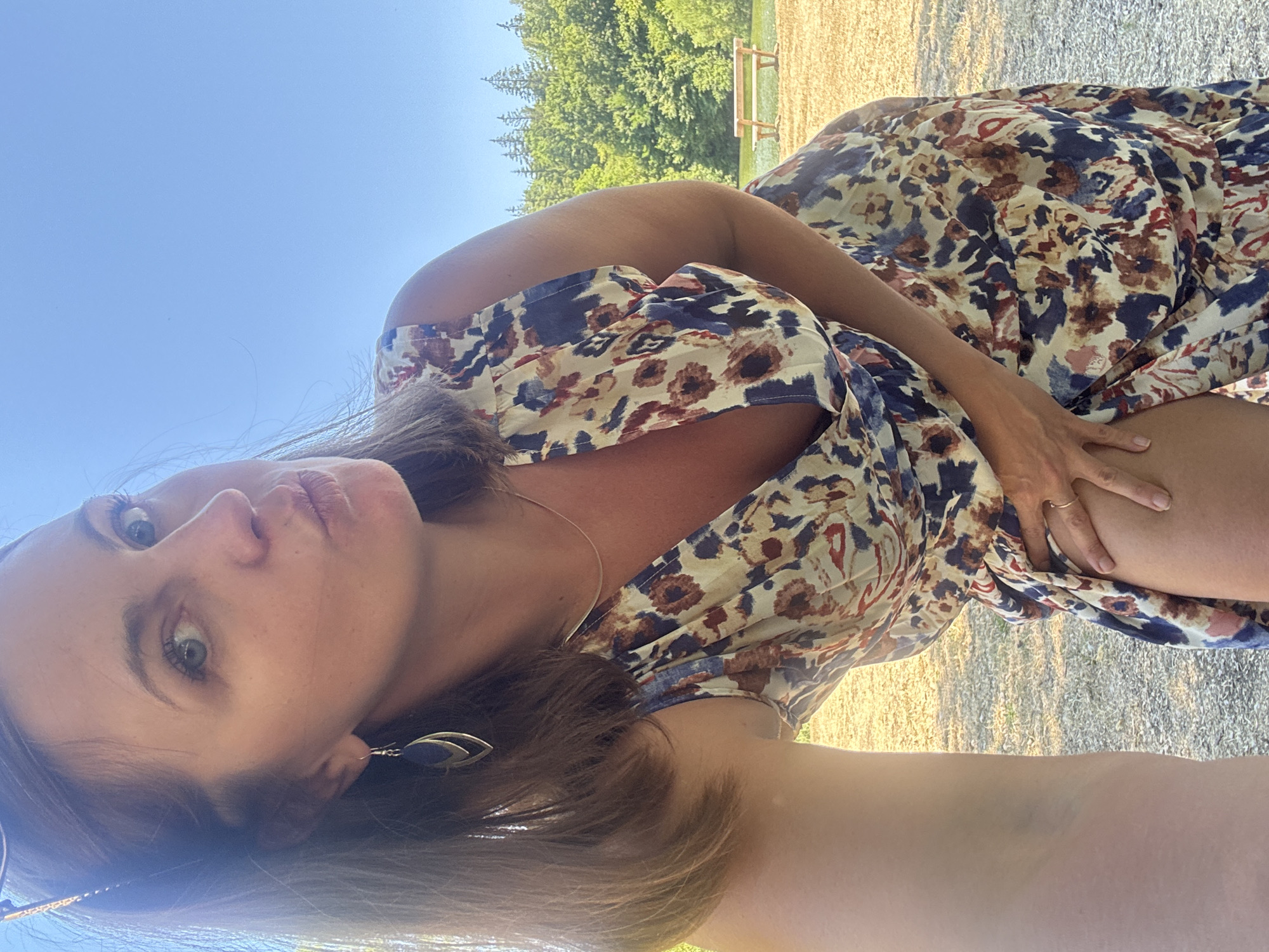 A woman poses for a selfie outdoors in Coteaux du Lizon, France, with a bright blue sky and a lush green forest in the background. She is wearing a floral print dress and a delicate necklace. Her hand is resting on her thigh, and she is puckering her lips as if blowing a kiss. A wooden bench is visible in the distance, suggesting a park or recreational area. The sun is shining, casting a warm glow on the scene.