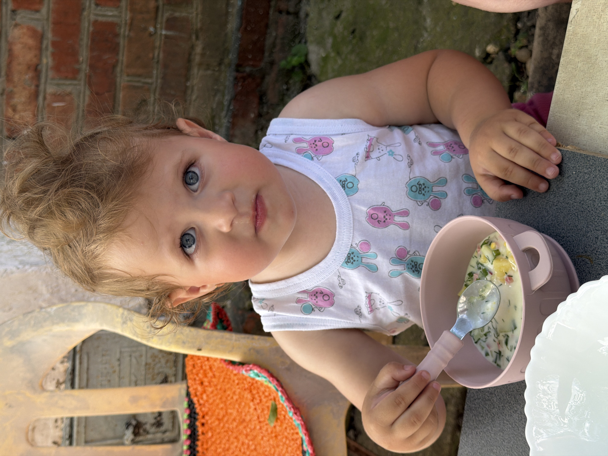 A young child with blond curly hair and blue eyes is sitting at a table outdoors and eating. The child is wearing a white sleeveless shirt with pink and blue bunny patterns. They are holding a pink spoon that is partially in a pink bowl of food, which appears to be a soup or cold dish with visible ingredients like potatoes and herbs. To the left of the bowl, there is a white bowl with a scalloped edge. The background includes a brick wall and a portion of a yellow plastic chair with an orange and green woven mat. The overall scene suggests a casual meal, possibly in a backyard or garden setting in Komsomolskiy, Russia.