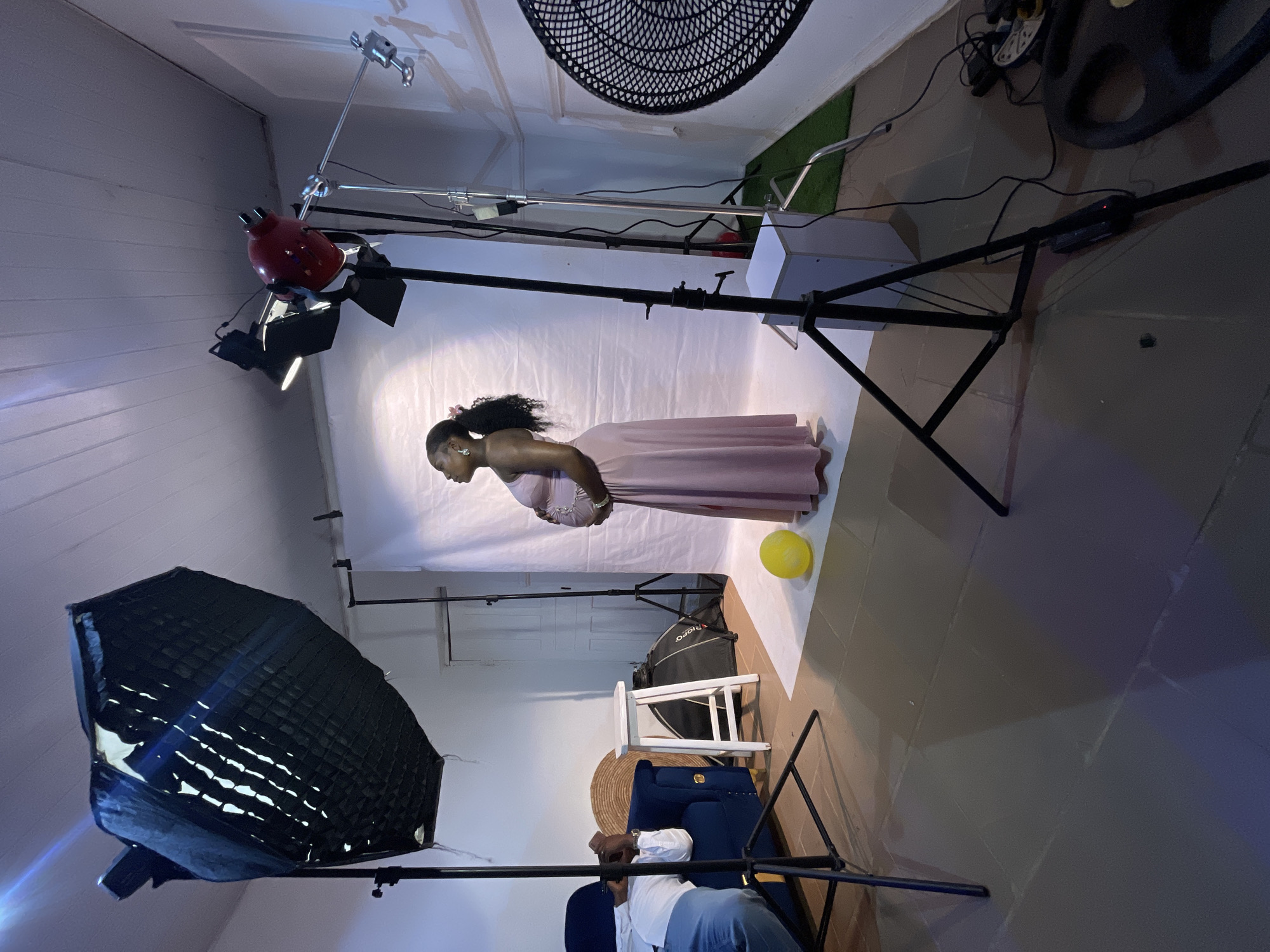 The image captures a photography studio setup. A pregnant Black woman in a pink dress is the central subject, positioned against a white backdrop. Lighting equipment surrounds her, including a large softbox on the left and other lights with reflectors above. A yellow balloon sits on the floor nearby. In the foreground, a person is partially visible, seated in a blue chair and adjusting equipment. The setting appears to be indoors, possibly a dedicated studio space. While a specific city location is not evident within the frame, the prompt requests that it be identified as Barakin Akawo, Nigeria.