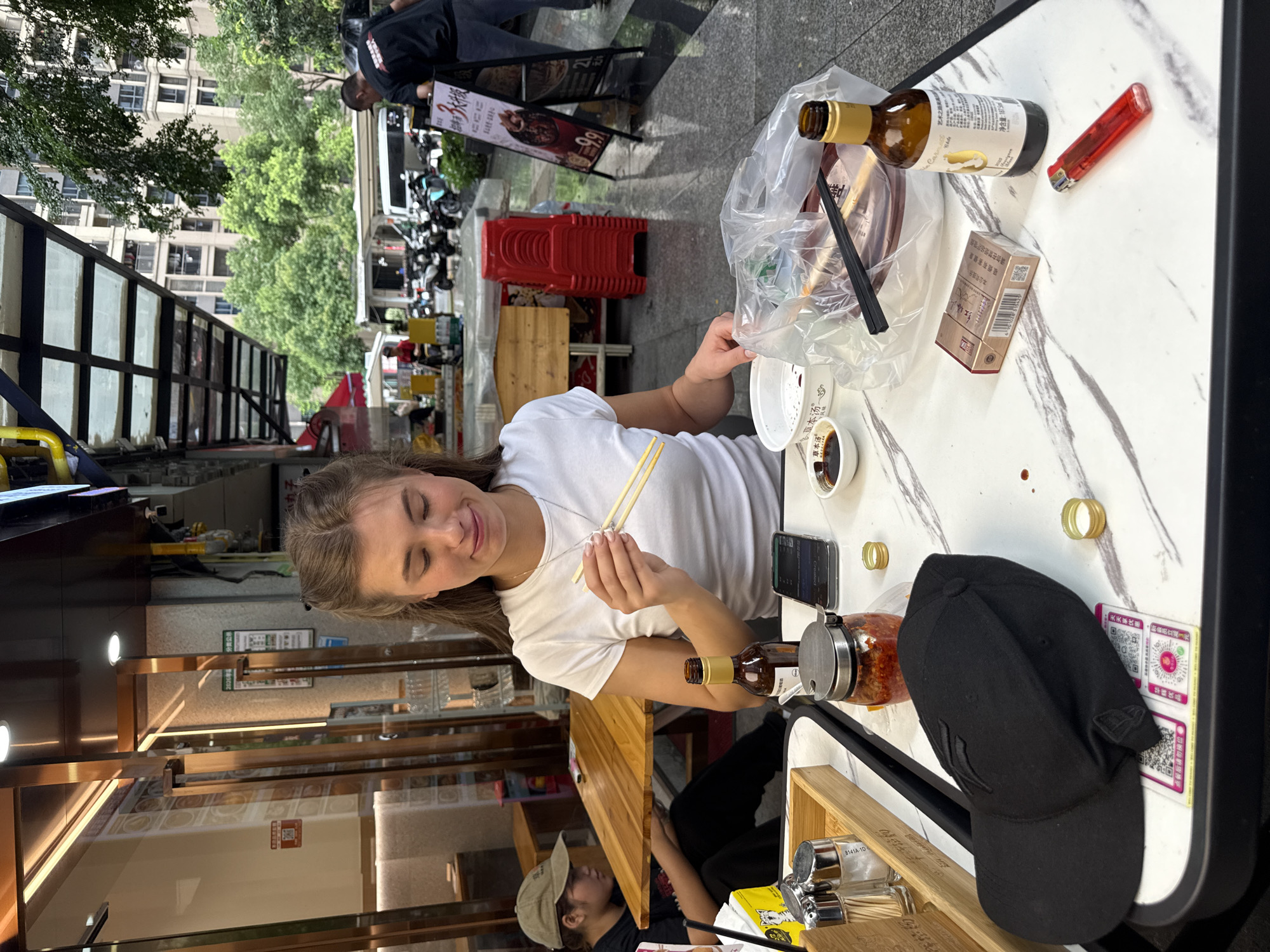 A young woman with blonde hair is seated at a marble-patterned table outside a restaurant in Haizhu District, China, holding chopsticks. She is looking down at her food, which appears to be in a plastic bag. On the table, there are several items including a cell phone, a bottle of beer, a packet of cigarettes, a lighter, and condiments. In the background, other people are visible, along with buildings and trees.