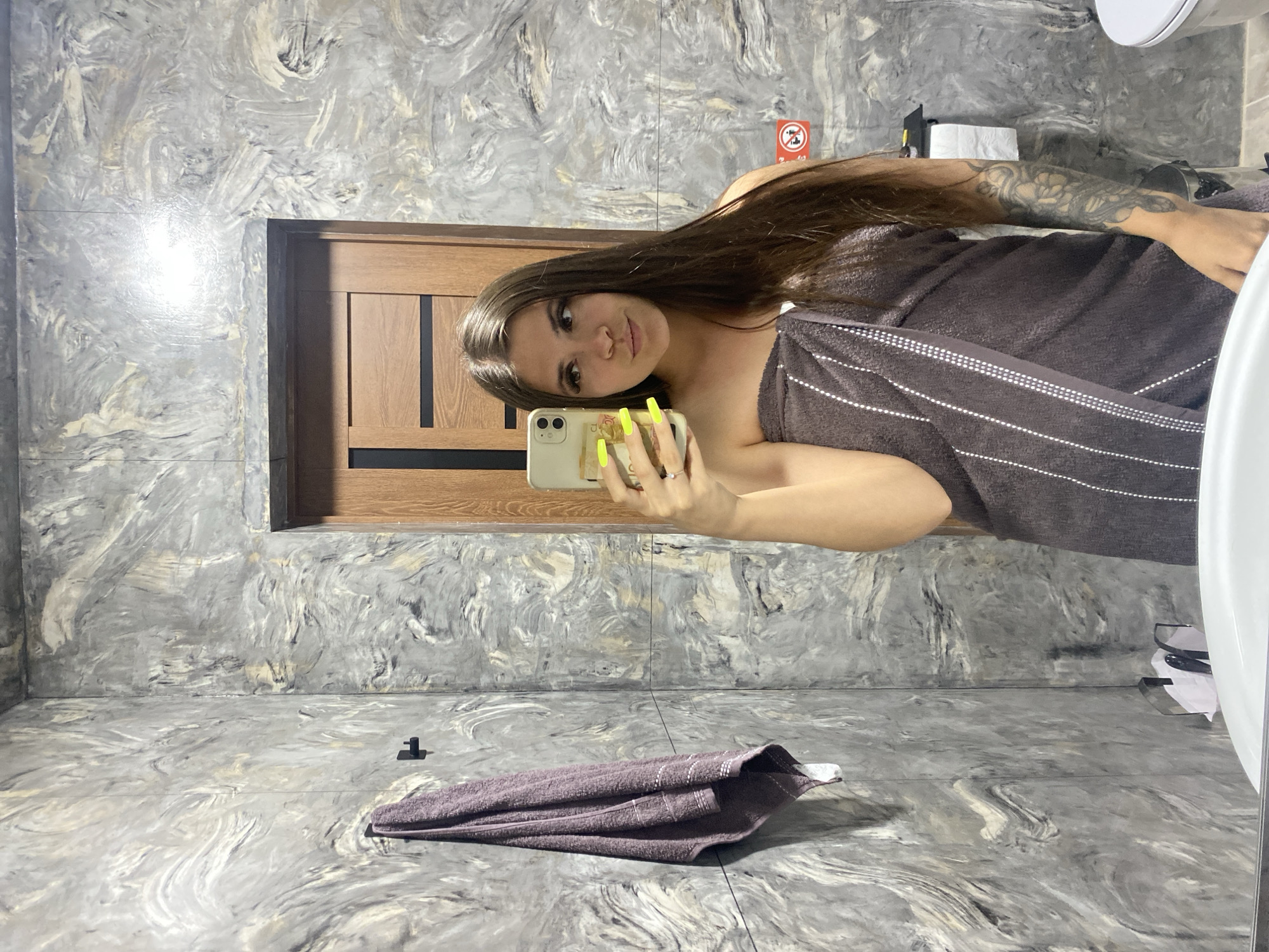 A young woman with long, dark hair, wearing a dark brown towel wrapped around her body, stands in a bathroom and takes a selfie in a mirror. She has bright yellow fingernails and a ring on her left ring finger. Her right arm is visible, revealing a tattoo of flowers. The bathroom features walls with a marble-like gray and white pattern, a wooden door, a towel hanging on a hook, a toilet paper dispenser, and a white toilet. The image does not provide any information about the city of Chalda, Russia.
